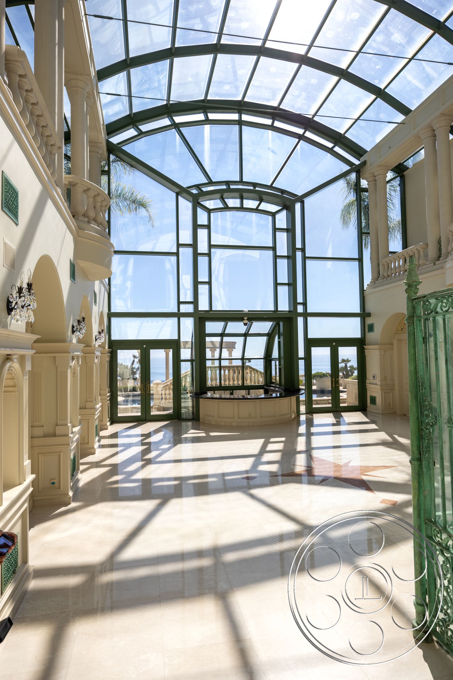 Pacifica Estate - resort, marble flooring, luxurious entrance hall, colonial architectural style, sun-drenched space, coastal view setting, open layout interior, natural light flooding interior, glass atrium ceiling, ornate column details, intricate wrought iron railings, arched glass windows, green metal framework, tall ceiling with skylight, elevated standing area, interior garden feel, polished marble entrance, elegant glass doors