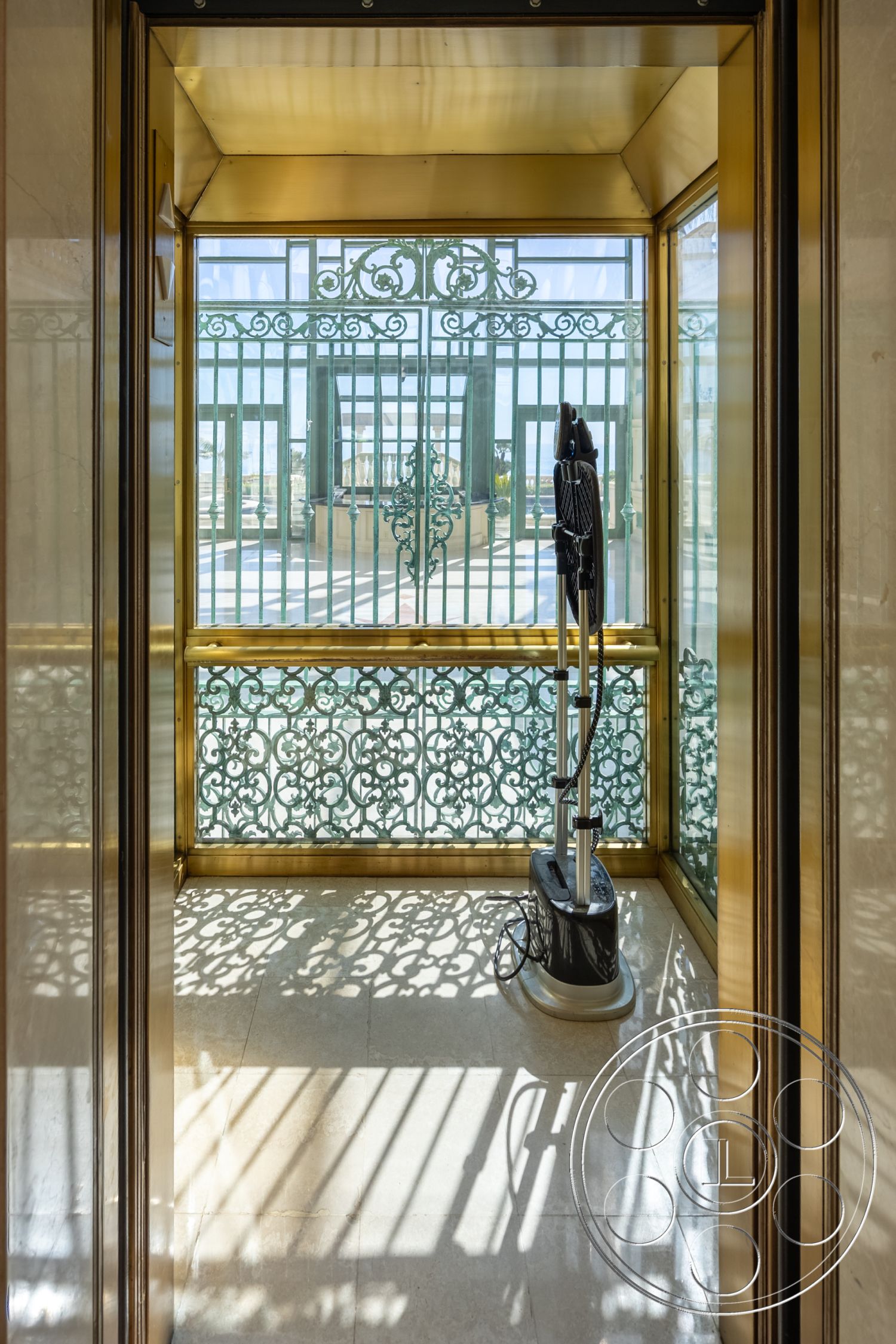 Pacifica Estate - high ceiling, polished marble flooring, scenic outdoor view, elegant architectural details, ornate wrought iron railing, decorative metalwork, ambient shadows, modern elevator interior, sleek modern aesthetics, interior elevator space, brass paneling, glass elevator walls, sunlight filtering through railings, luxury elevator design, commercial-grade elevator