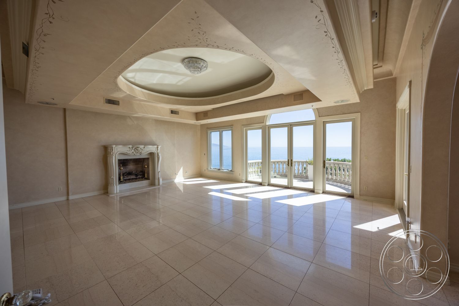 Pacifica Estate - spacious layout, coffered ceiling, open floor plan, sliding glass doors, floor-to-ceiling windows, ocean view, marble flooring, arched doorways, luxurious living room, elegant chandelier, natural light-filled space, traditional fireplace surround, decorative ceiling molding, soft beige wall treatment