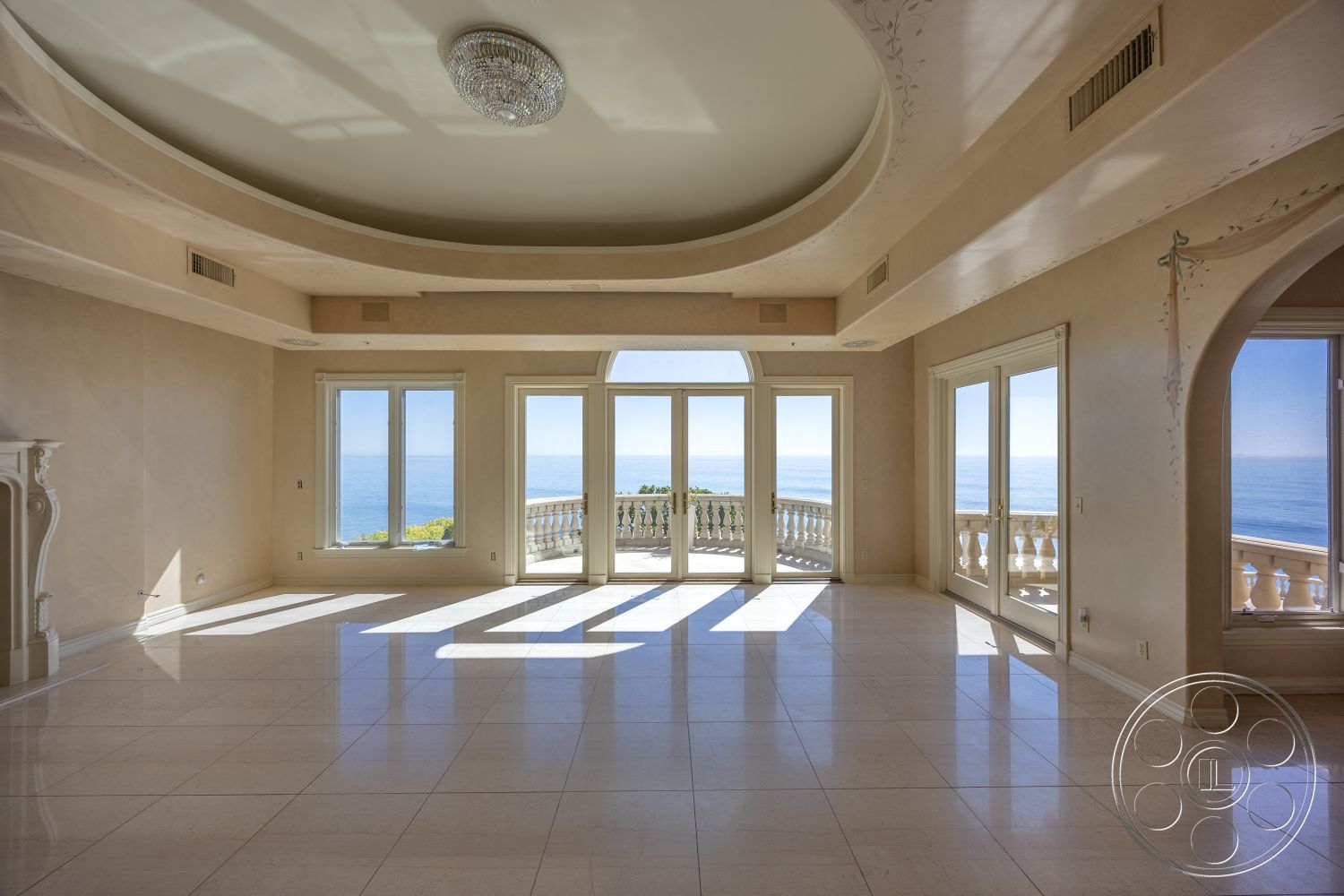 Pacifica Estate - neutral painted walls, elegant chandelier lighting, interior marble floor tiles, arched ceiling with crown molding details, expansive glass windows with ocean view, exposed columns at entry, balcony with decorative balustrade, light-filled open floor plan, large glass sliding doors to exterior terrace, natural light reflection on polished surfaces, coastal contemporary design, luxurious living space with panoramic views