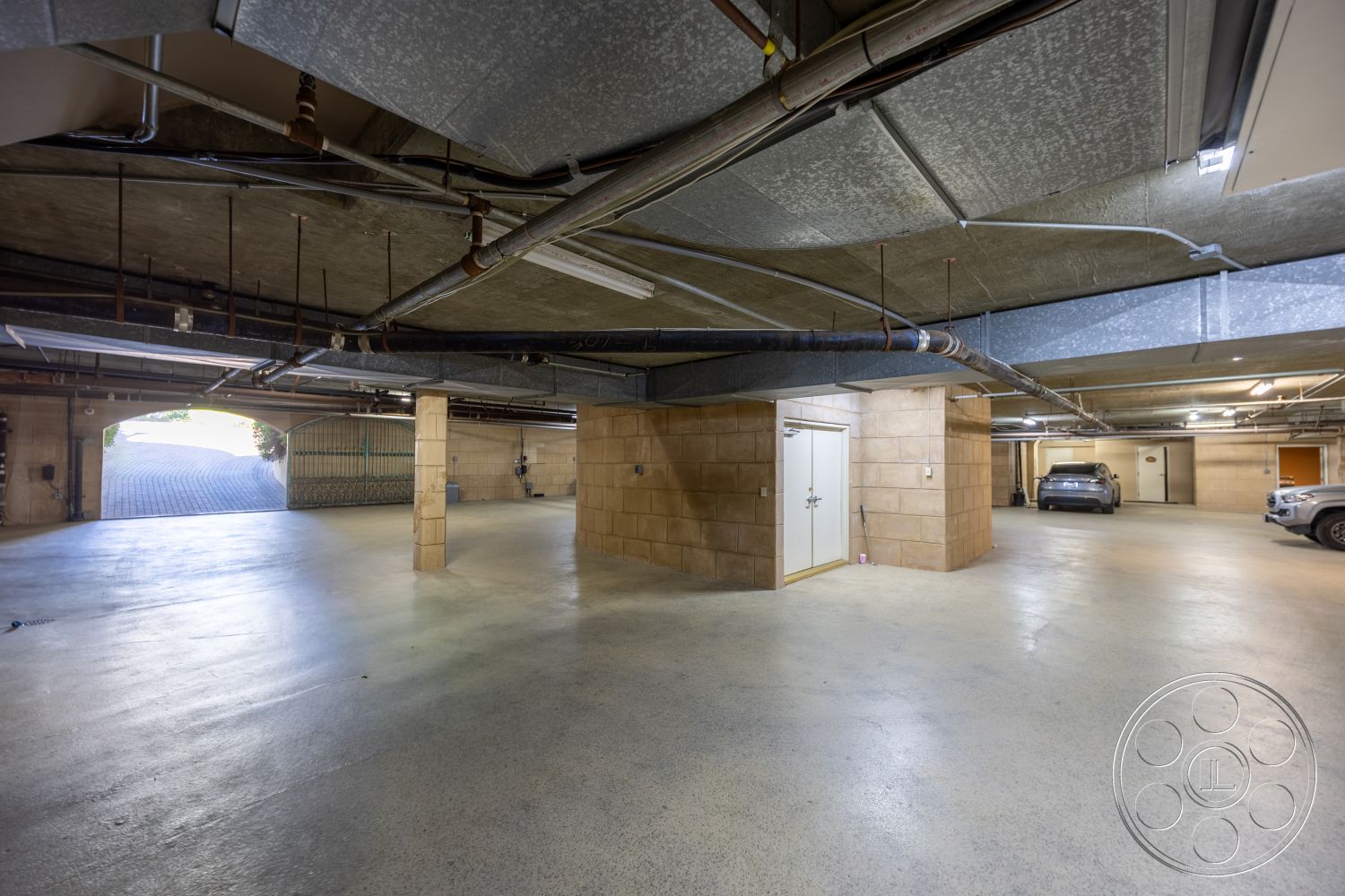 Pacifica Estate - concrete flooring, smooth concrete surface, industrial lighting fixtures, brick accent wall, column structure, circular layout, painted walls, natural light entry, interior garage space, exposed ceiling with pipes, utility room door, paved driveway access, central support columns