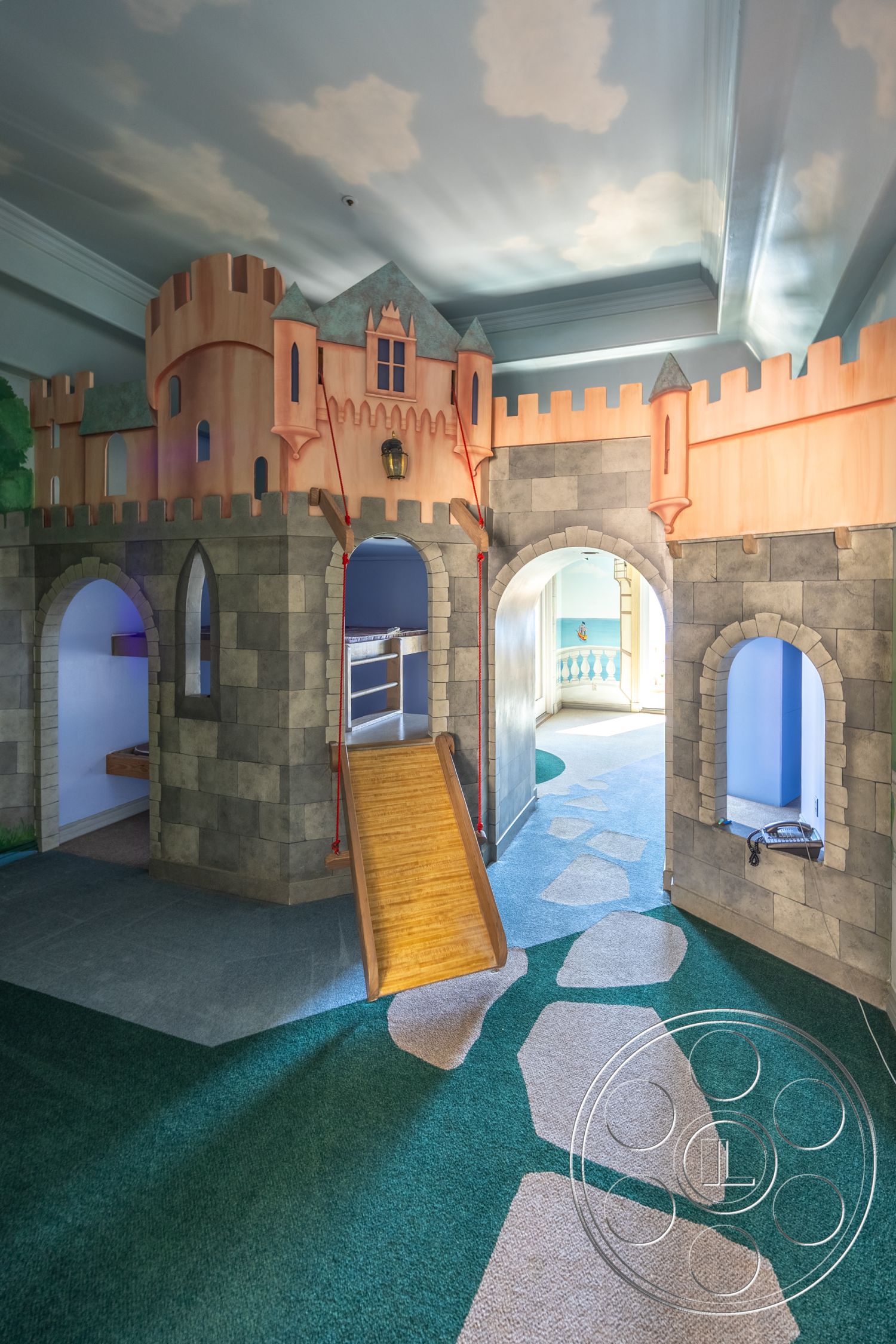 Pacifica Estate - textured plaster walls, arched entryways, green indoor carpet flooring, interior castle-themed play area, stone façade walls, wood slide ramp, playhouse with turrets and battlements, blue ceiling with cloud murals, open play layout, decorative lantern fixtures