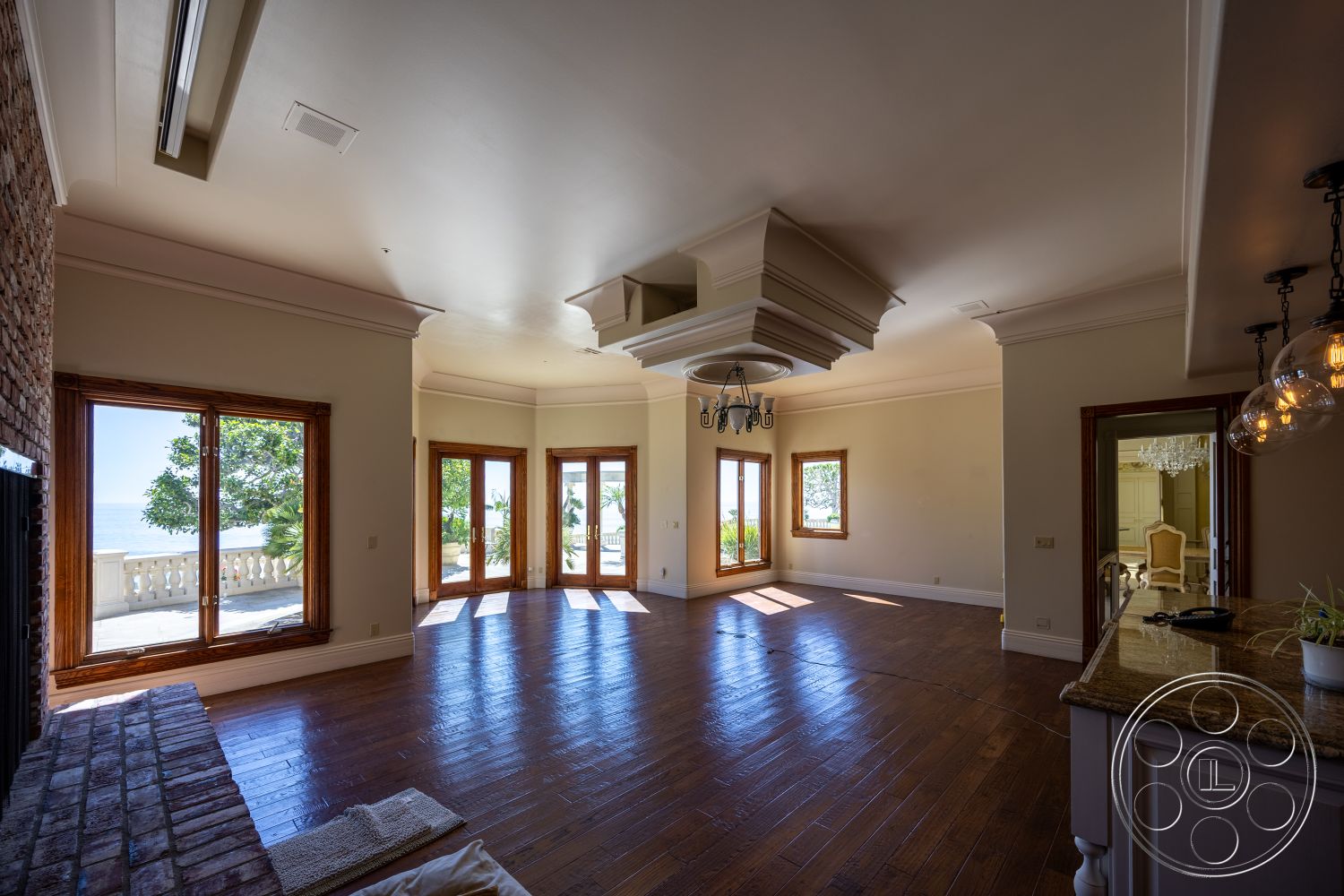 Pacifica Estate - spacious layout, coffered ceiling, vaulted ceiling, elegant lighting fixtures, subdued color palette, beachfront property, granite kitchen countertop, hardwood living room floors, exposed brick fireplace surround, crown molding details, interior space with natural light, open living area, double glass door entry, large windows with ocean views, decorative drop ceiling, coastal interior design, detailed wood trim, elevated ceiling placement