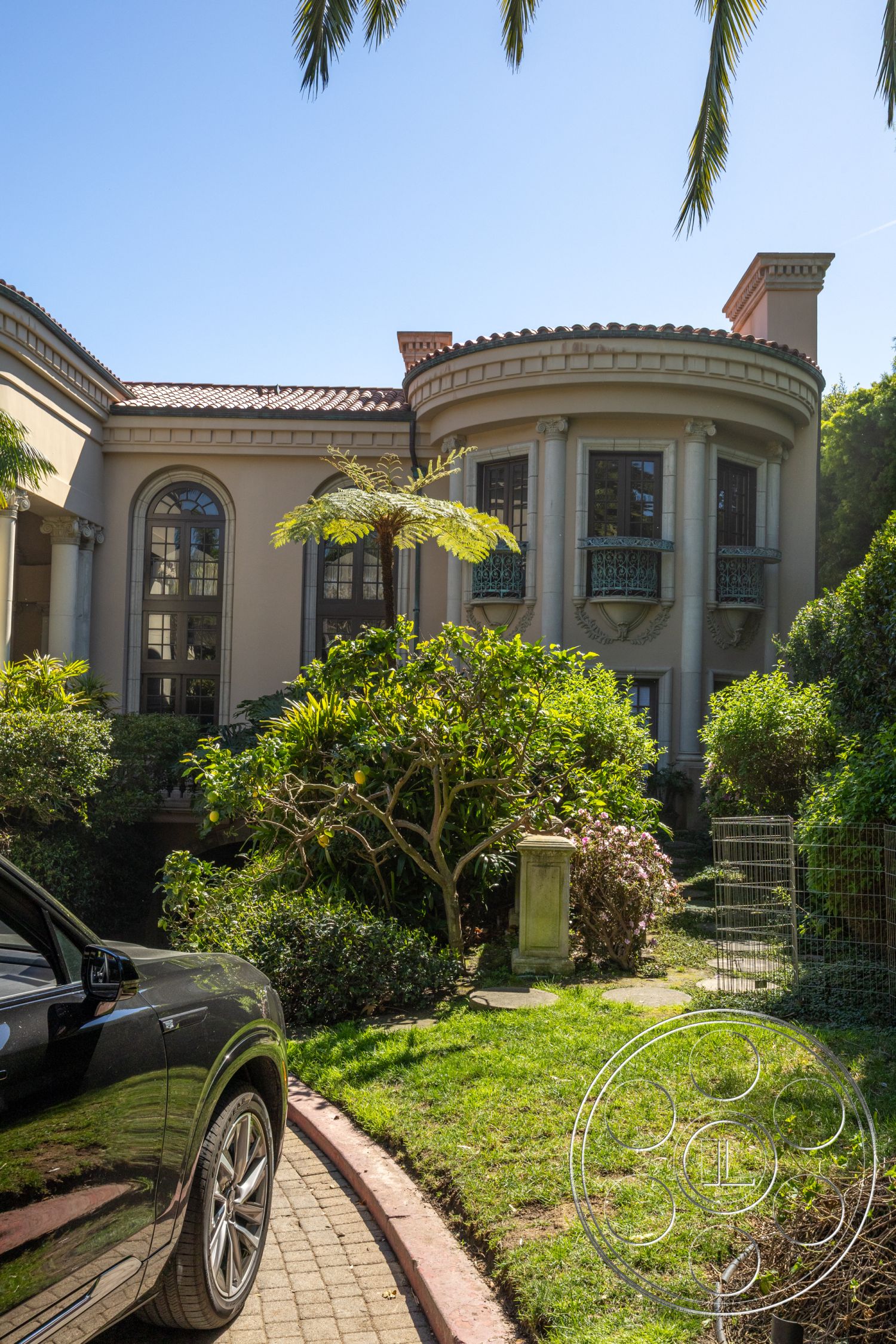 Pacifica Estate - lush tropical landscaping, green manicured lawn, stucco exterior walls, lush garden setting, sunny outdoor environment, tile roof with red clay tiles, curved balcony with wrought iron railings, circular driveway with interlocking pavers, arched windows with decorative moldings, ornamental column details, front patio with stone pathway, exterior with Mediterranean architectural style, front yard with palm trees, exposed beam ceilings visible from exterior, historic villa aesthetics