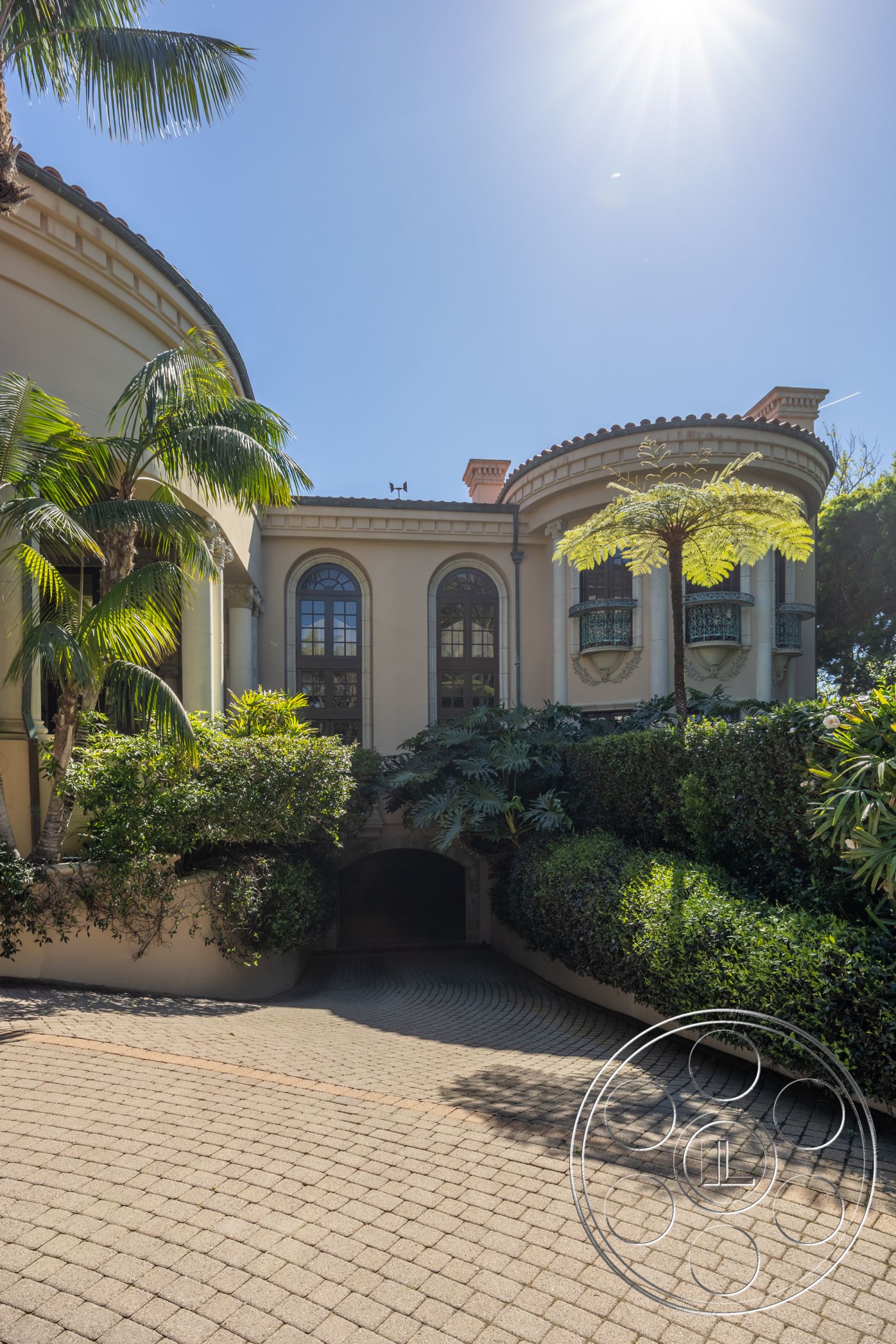 Pacifica Estate - paved driveway, landscaped greenery, circular driveway, gated entrance, arched doorways, cobblestone path, tropical setting, hillside property, two-story house, Mediterranean architectural style, sunlit exterior, stucco exterior walls, exterior palm trees, curved architectural forms, balcony railings, ornate window details, plant-filled entry, stone facade accents, garage access, luxurious home exterior