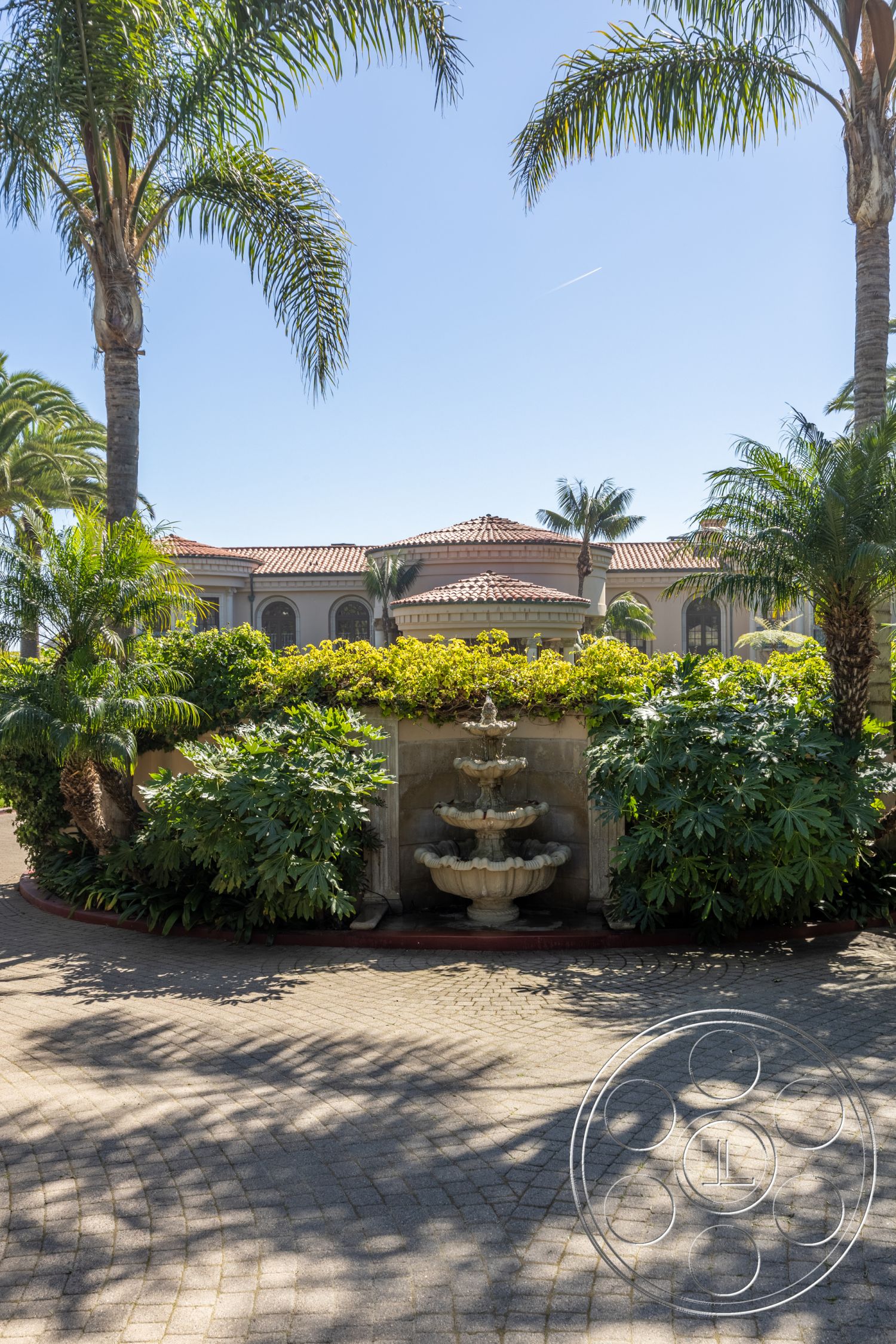 Pacifica Estate - exterior, palm trees, paver driveway, outdoor living space, tropical landscaping, colorful flowering plants, decorative stone wall, mediterranean architecture, lush green foliage, urban oasis setting, brick driveway border, rounded fountain, stone fountain detail, arced fountain structure, sunlit garden patio, curved driveway edge