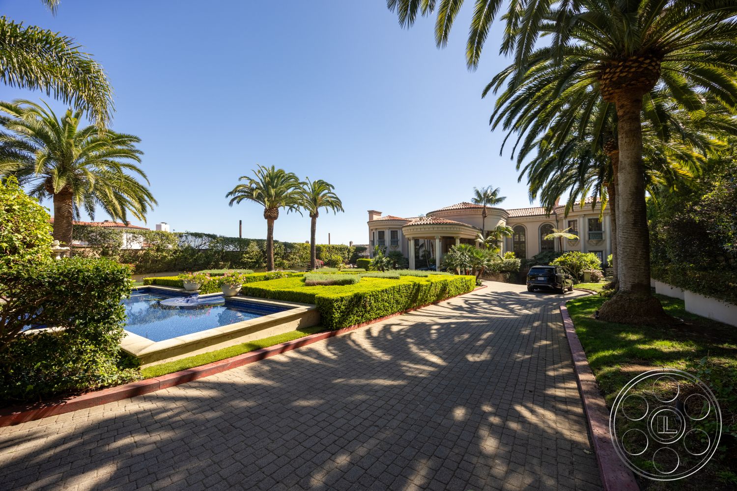 Pacifica Estate - sunny coastal setting, concrete pool deck, Mediterranean architectural style, brick driveway pavers, outdoor patio area, stone pool border, exterior landscaping with palm trees, surrounding hillside views, rectangular swimming pool with water feature, lush garden greenery, multicolored flower beds, spacious front lawn with hedges, sloped driveway with curvature, covered entrance porch, large windows with arched details, fountain feature in swimming pool, palm-fringed garden areas