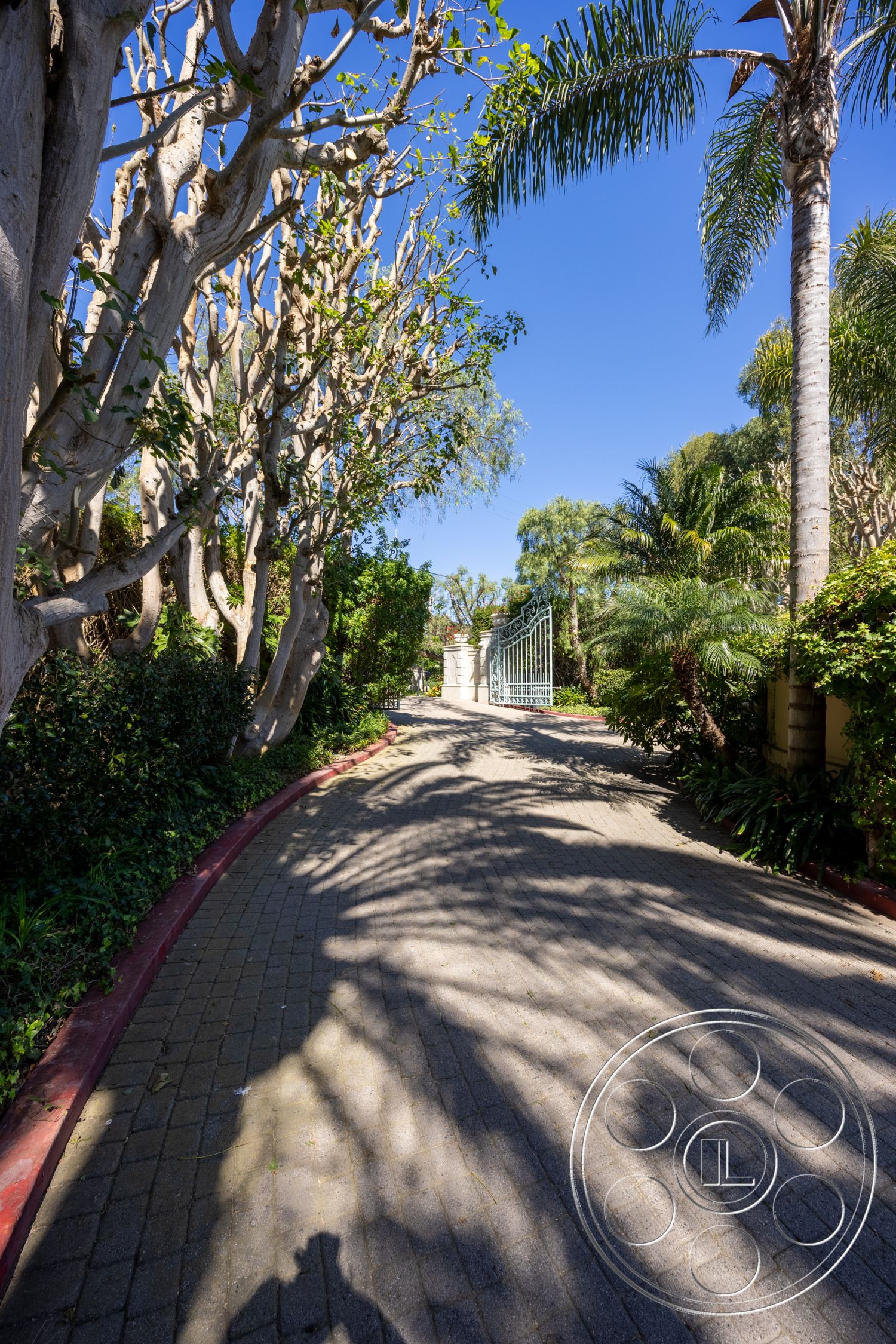 Pacifica Estate - exterior, landscaped garden, paved driveway, lush greenery, curved driveway, brick border, tropical palm trees, suburban backyard, private driveway, sunlit pathway, metal gate entrance, stone driveway pavers, shadow play on pavement, arched gateway
