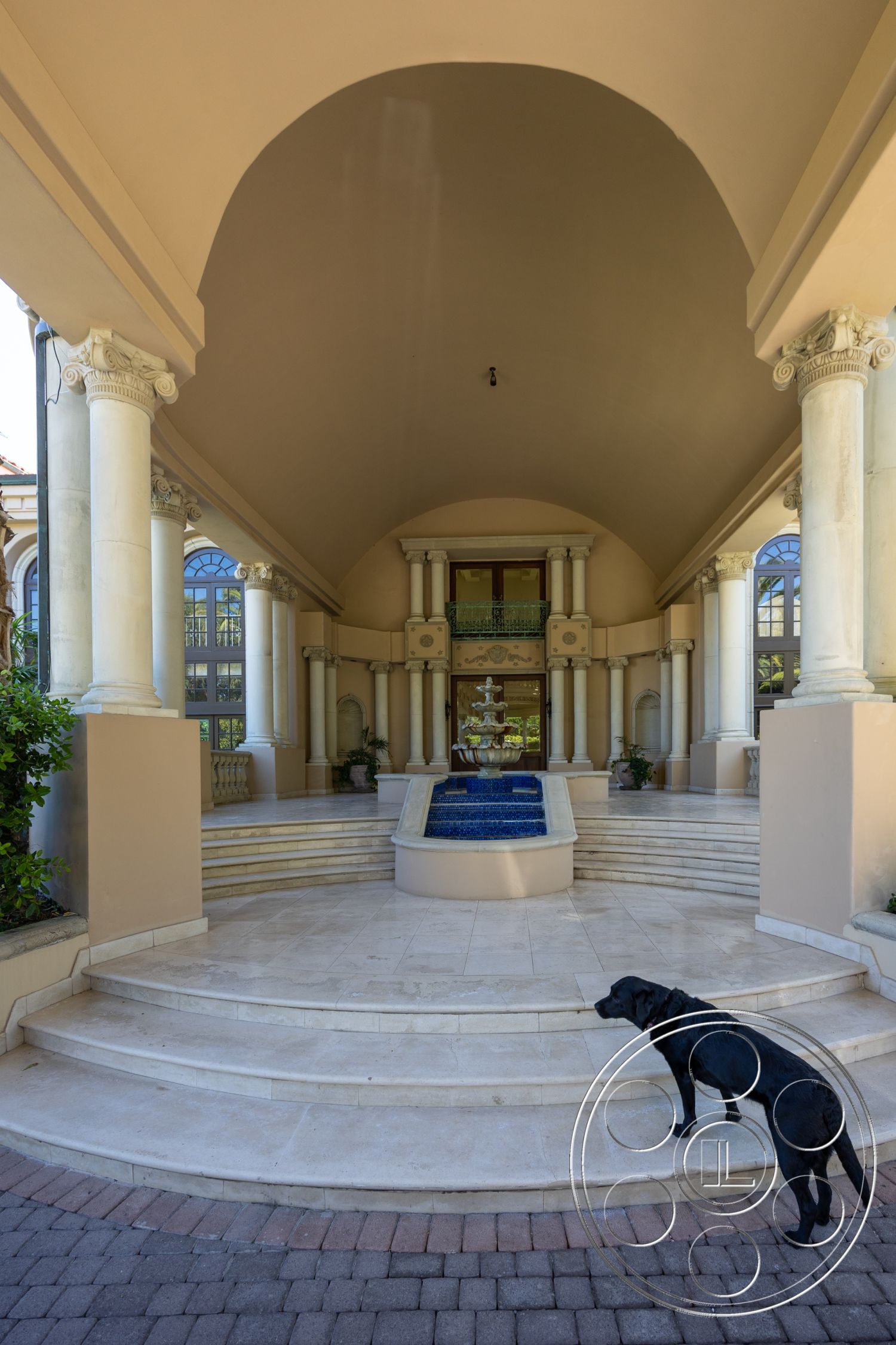 Pacifica Estate - textured plaster wall finish, decorative wrought iron balcony railing, surrounding garden landscape, open-air courtyard setting, exterior entrance with arched ceiling, marble steps leading to a fountain, stone column supports, large glass windows with decorative trim, paver stone patio flooring, water feature with blue tile accents, landscape with planters on either side, symmetrical stairway design, classical architectural elements, luxurious outdoor entrance area, curved entry stairwell