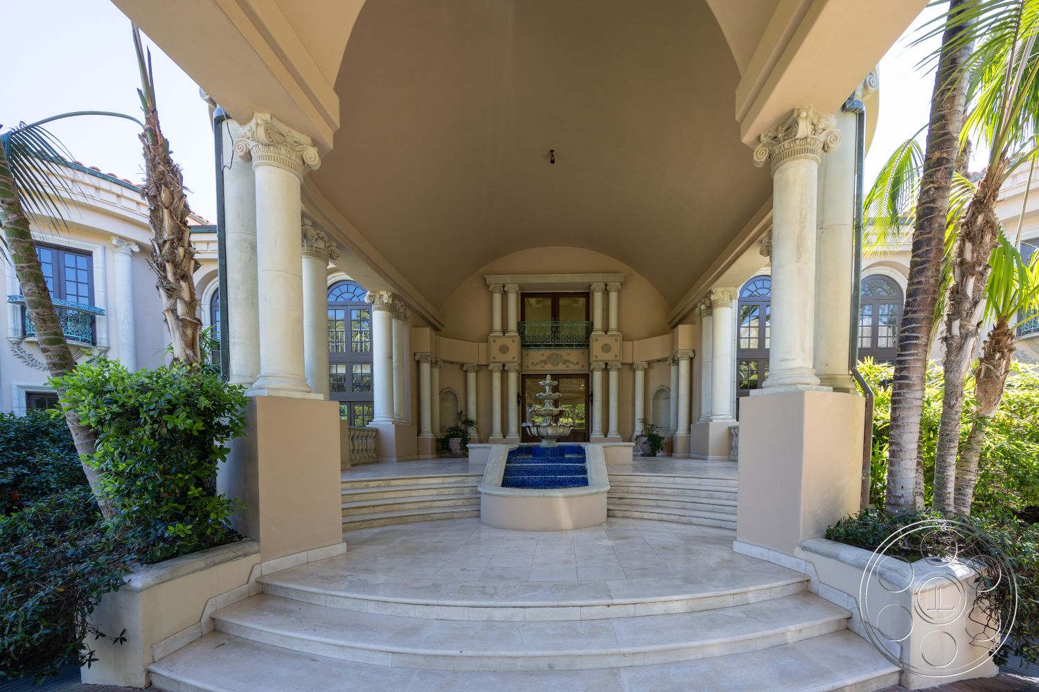 Pacifica Estate - natural light, landscaped garden, high ceiling, planted greenery, symmetrical design, arched ceiling, textured stone walls, decorative stone planters, exterior entrance, glass double doors, outdoor patio area, courtyard style, tranquil water feature, marble steps, columned porch with decorative capitals, blue fountain feature, palm trees flanking entrance, luxurious Mediterranean style, detailed wrought iron balcony, stone exterior facade