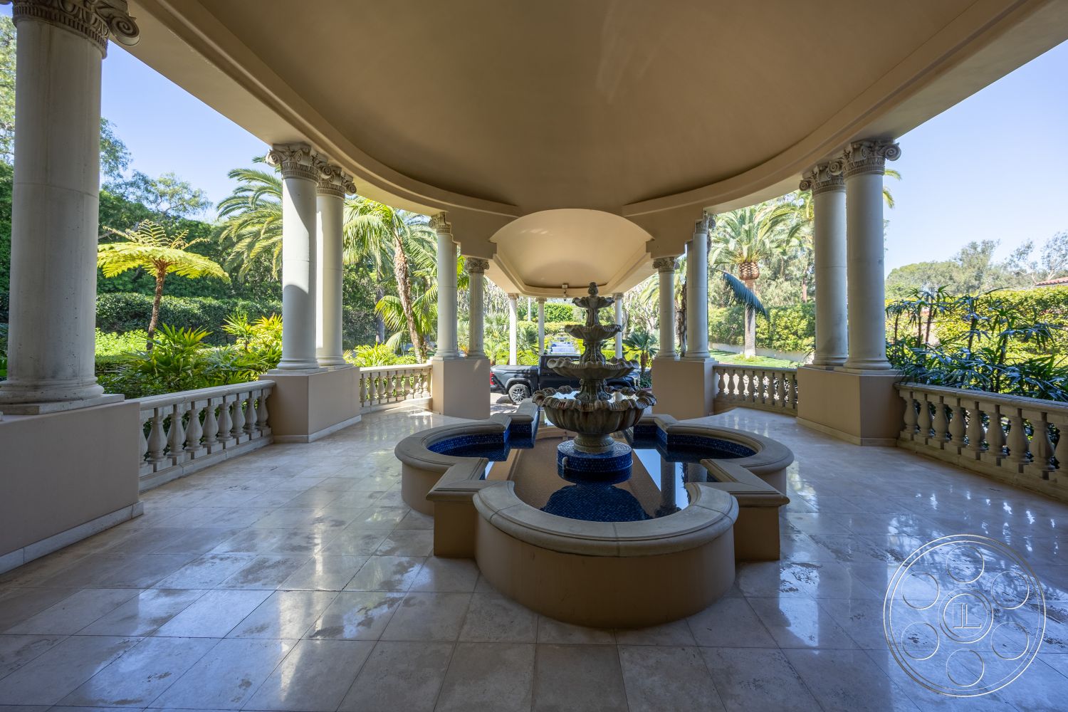 Pacifica Estate - landscaped garden with tropical plants, exterior colonnade with classical columns, marble flooring with polished finish, ornate stone fountain with water feature, balustrade railing with decorative details, arched ceiling with decorative molding, outdoor sitting area with circular seating, glistening water feature surrounded by greenery, paved entrance with unobstructed view, elegant patio area with tile accents, lush garden setting with palm trees, luxurious exterior design in Mediterranean style, spacious open air porch with natural lighting, custom-designed outdoor space for entertainment