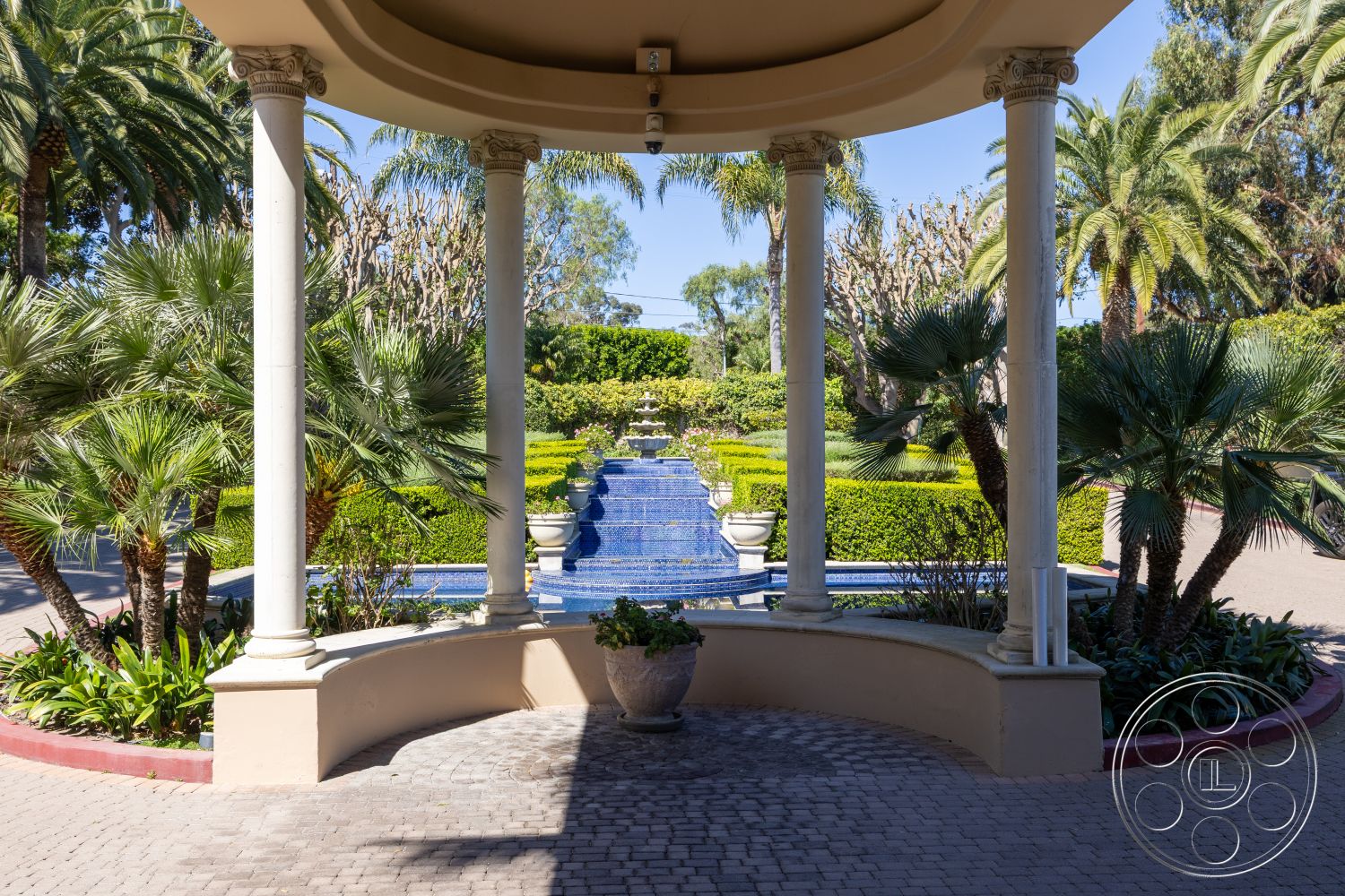 Pacifica Estate - tropical plants, landscaped greenery, stone pathway, brick pavers, fountain feature, arched entrance, circular layout, scenic backdrop, view of garden, reflected water surface, formal garden setting, ornamental hedges, sunlit outdoor space, mediterranean architectural style, terracotta flower pots, concrete bench seating, curved patio area, exterior columns, blue mosaic tiles, palm trees surrounding
