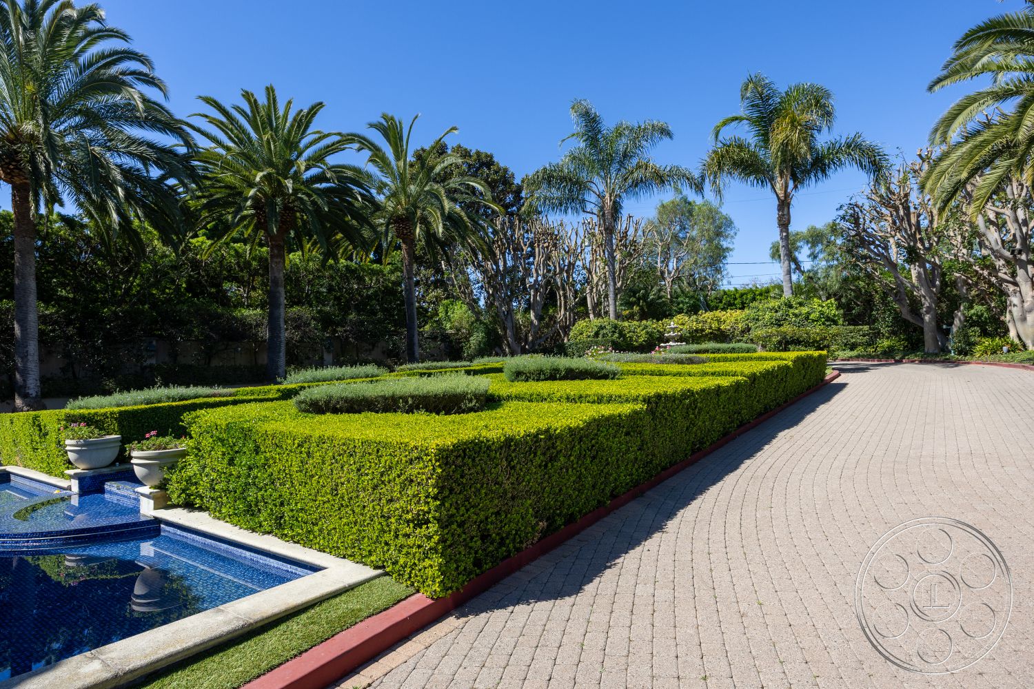 Pacifica Estate - curved walkway, bright blue sky, suburban backyard oasis, open green lawn area, rectangular swimming pool with mosaic tile, paved circular driveway, interlocking brick driveway pavers, surrounding tropical palm trees, manicured hedges with geometric shapes, stone pool deck with coping, luxurious landscape design, garden area with flower beds, exterior Mediterranean-style home, tropical outdoor setting, flower pots with blooming plants, textured concrete edging, drought-resistant landscaping features