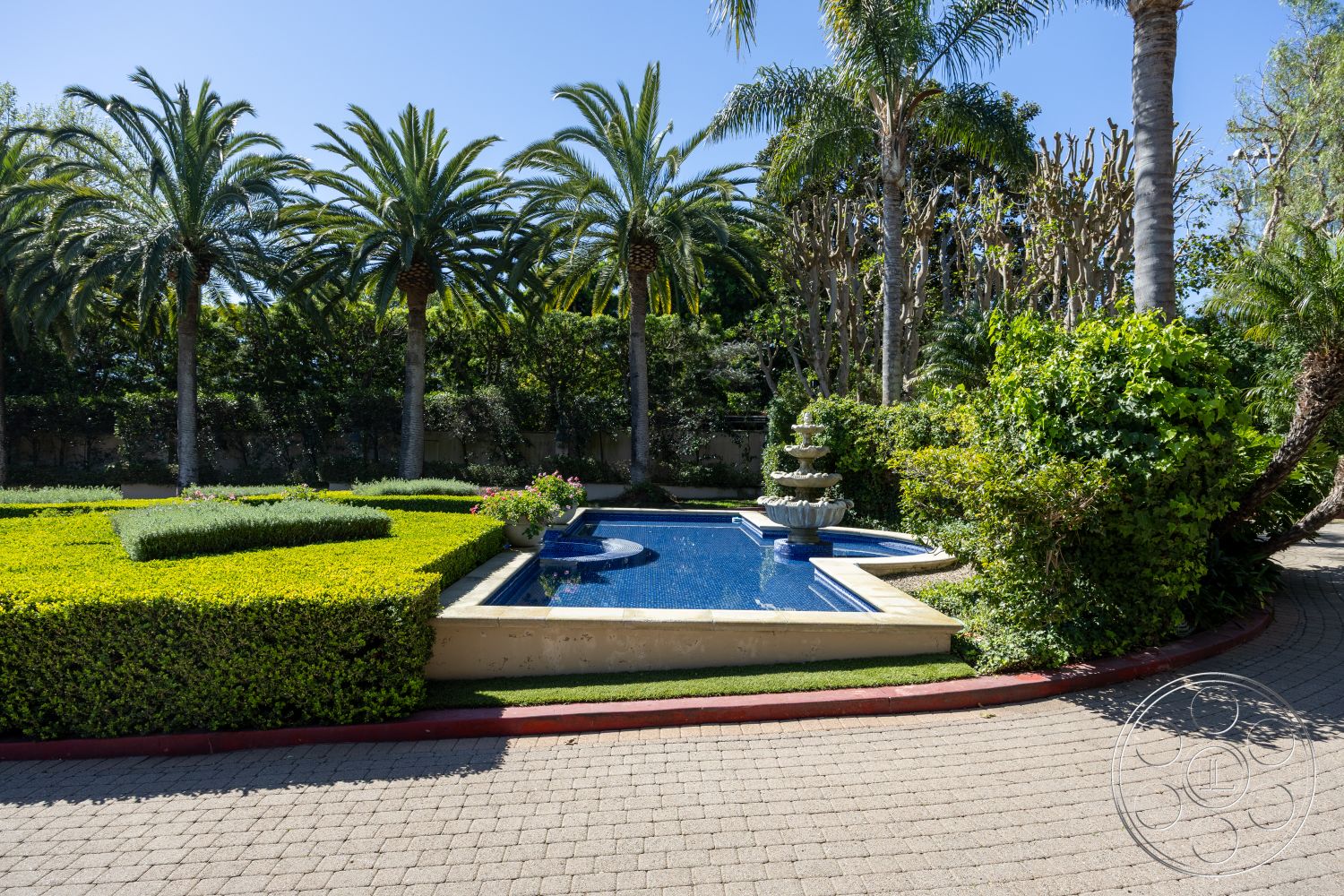 Pacifica Estate - exterior, rectangular swimming pool, urban oasis, concrete pool deck, modern outdoor space, lush greenery backdrop, luxurious residential property, paved stone driveway, textured concrete borders, blue ceramic pool tiles, landscape garden with manicured hedges, fountain feature in pool area, tropical palm trees surrounding pool, garden area with colorful flowers, hardscape with integrated greenery, inviting outdoor setting