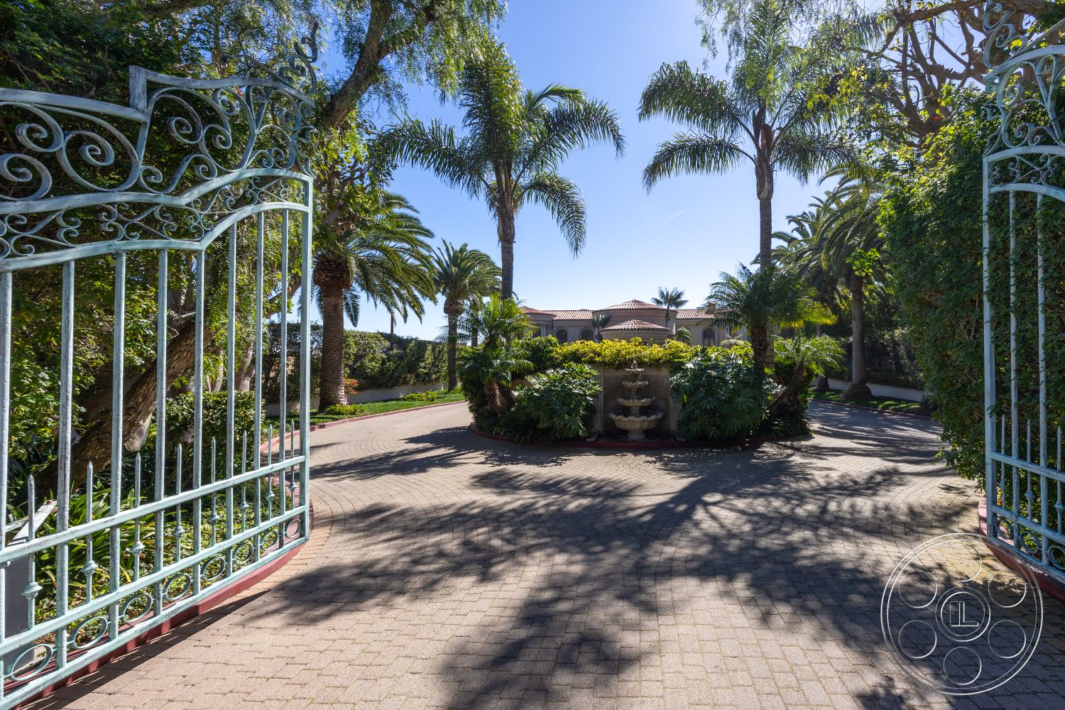 Pacifica Estate - lush tropical landscaping, residential exterior, sunny outdoor setting, textured stone fountain, desert climate setting, surrounding hillside views, paver stone driveway, ornate wrought iron gate, paved circular driveway, palm trees lining the entrance, koi pond with fountain feature, gated estate entrance, mature greenery and shrubs, mediterranean style architecture, brick border garden pathway, irregular shaped garden beds, decorative landscape lighting