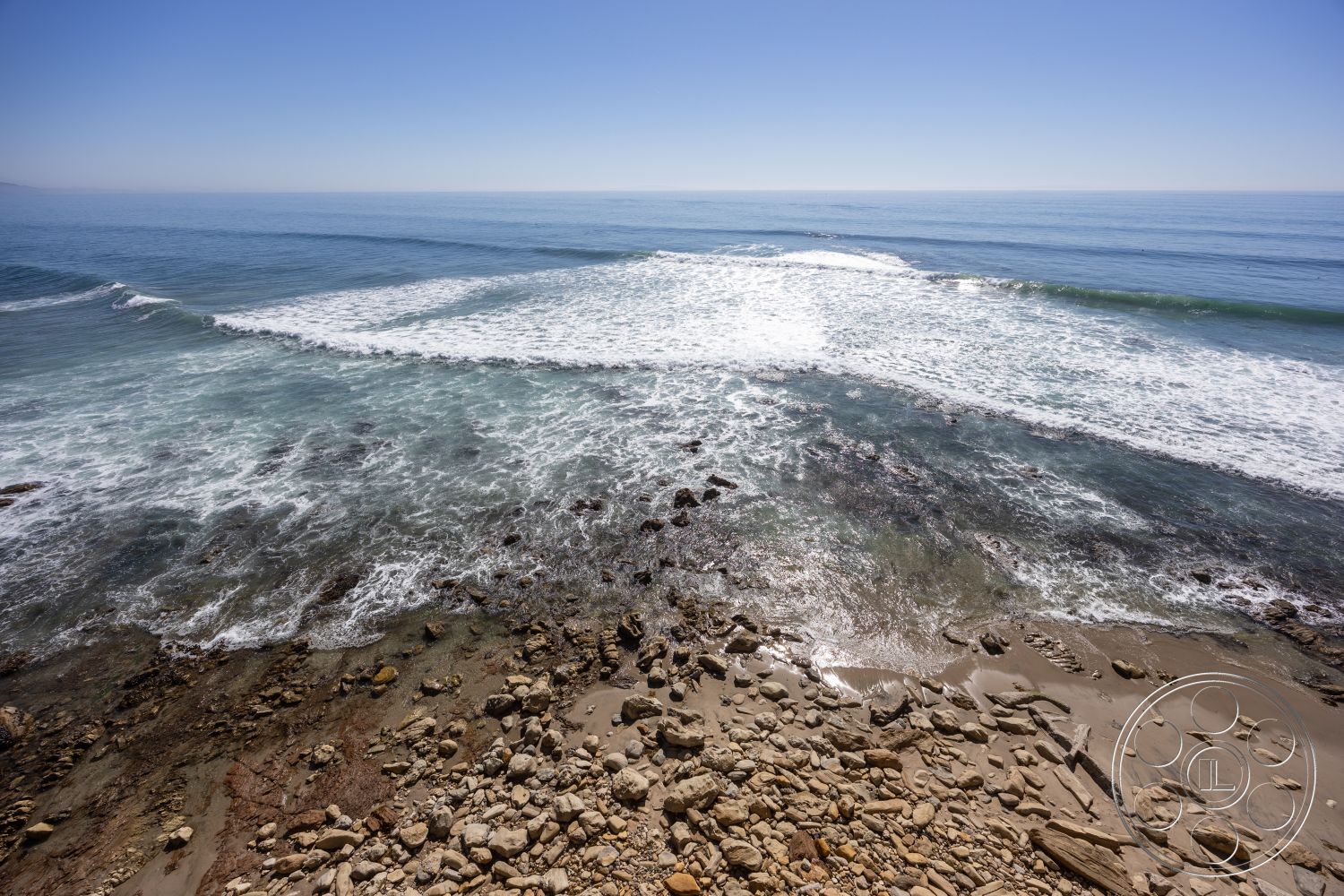 Pacifica Estate - clear blue sky, natural stone textures, coastal environment, exterior coastal view, beachfront setting, rocky beach shoreline, sandy beach area, ocean waves crashing, tidal pool formations, scattered pebbles and stones, marine landscape, gentle ocean currents, sunlight reflecting on water, marine vegetation along shore, photo captured at midday