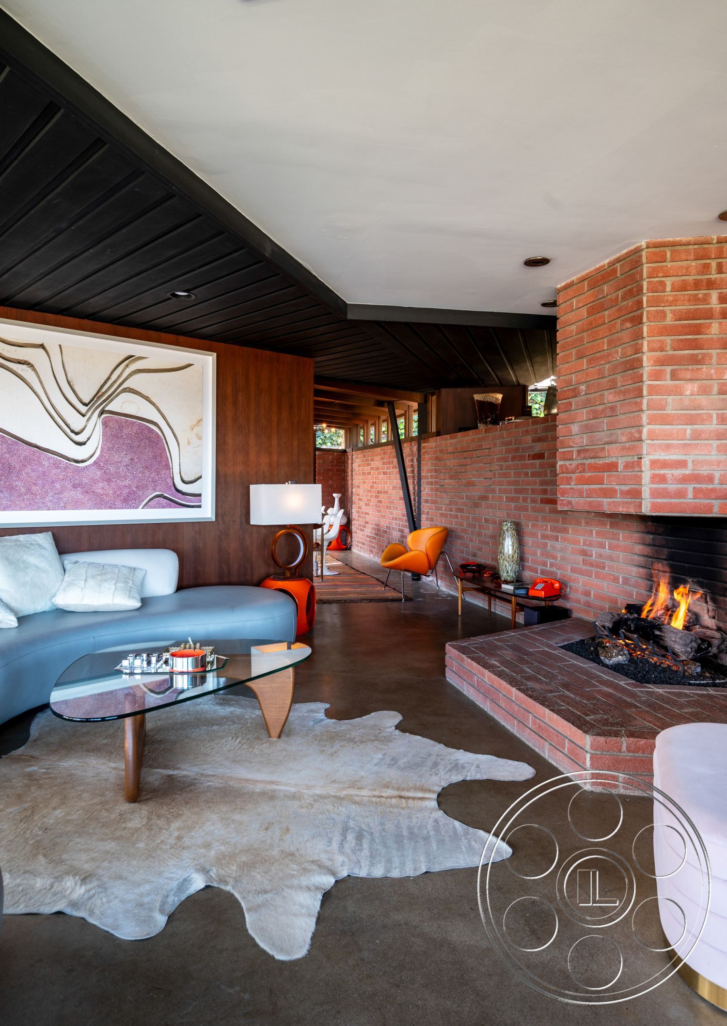 Retro 31 - minimalist decor, open floor plan, concrete floor, glass coffee table, pendant lighting, warm color palette, artwork on wall, exposed brick fireplace, brick accent wall, mid-century modern furniture, black wood beam ceiling, curved sectional sofa, cowhide area rug, colorful accent chair