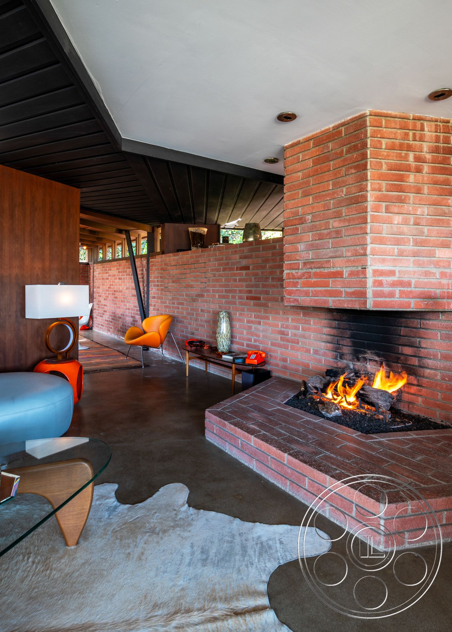 Retro 31 - floor-to-ceiling windows, industrial lighting fixtures, warm ambient lighting, textured plaster walls, mid-century modern furniture, modern minimalist decor, exposed brick fireplace surround, neutral color palette interior, open concept living area, concrete floor finish, cowhide area rug, black ceiling with wooden beams, glass coffee table with wooden legs, bright orange accent chairs, rustic wooden accent wall, cozy seating area with fireplace