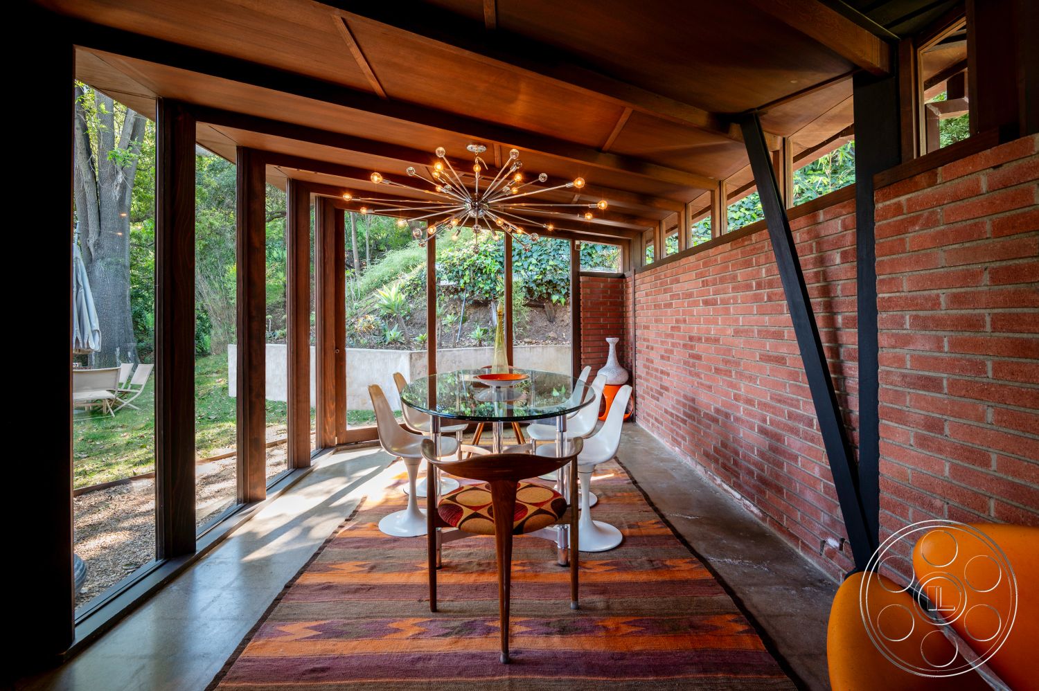 Retro 31 - mid-century modern architectural style, natural light interior, floor-to-ceiling windows interior, open floor plan interior, exposed brick accent wall interior, concrete floor interior, textured area rug interior, modern lighting fixture interior, hardwood ceiling beams interior, glass dining table interior, white modern dining chairs interior, orange accent chairs interior, lush garden view setting, wood frame structure interior, greenery surrounding setting