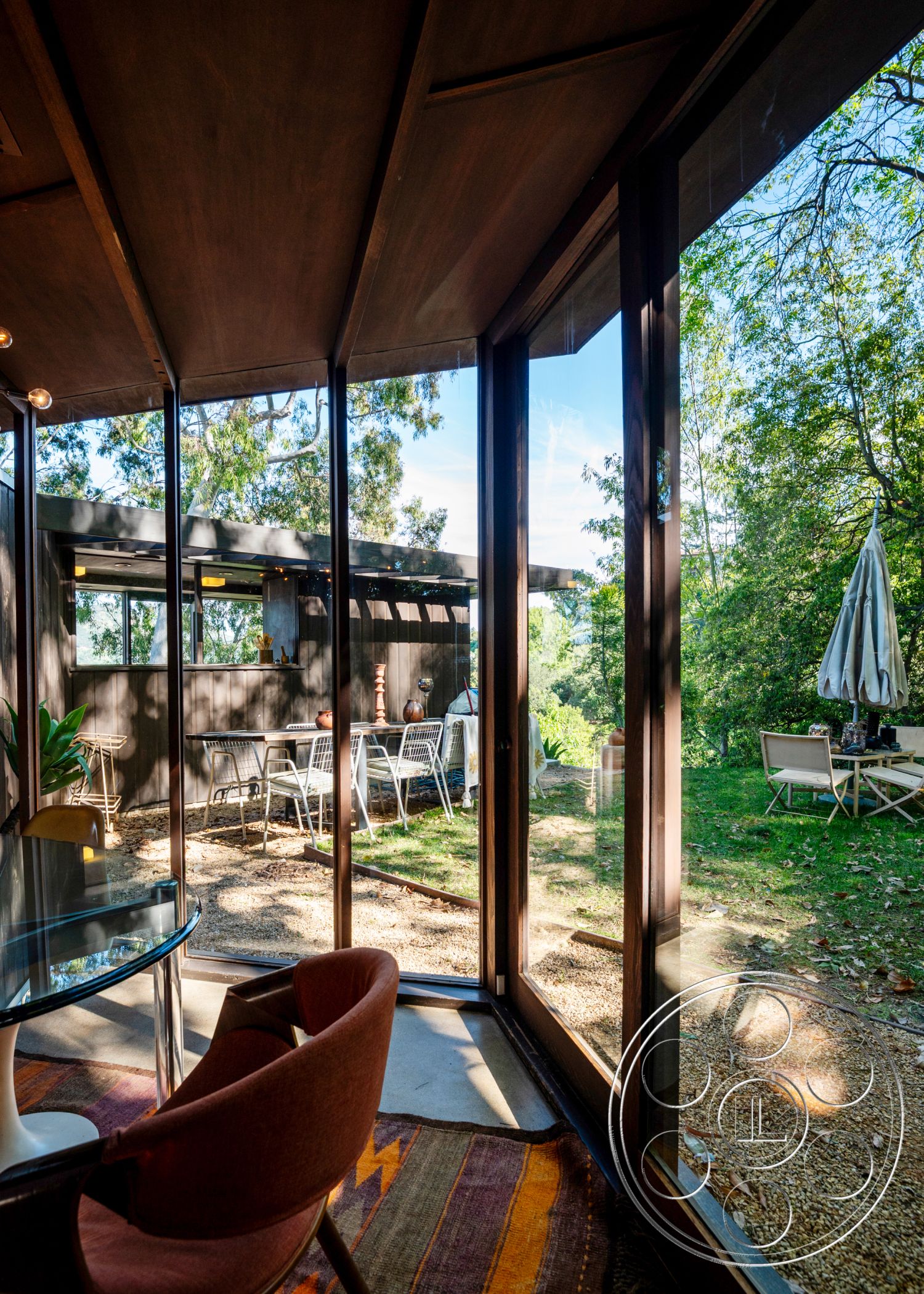 Retro 31 - glass sliding doors, metal outdoor furniture, gravel landscaping, seamless indoor-outdoor flow, textured wall treatment, modern dining area, curved roof design, landscaped garden setting, mid-century modern architectural style, natural light interior, overhanging roof eaves, outdoor dining patio, exterior glass walls, bright, airy atmosphere, elevated terrace area, interior wood ceiling structure, concrete floor with rug, surrounding tree coverage, dining table with wire chairs