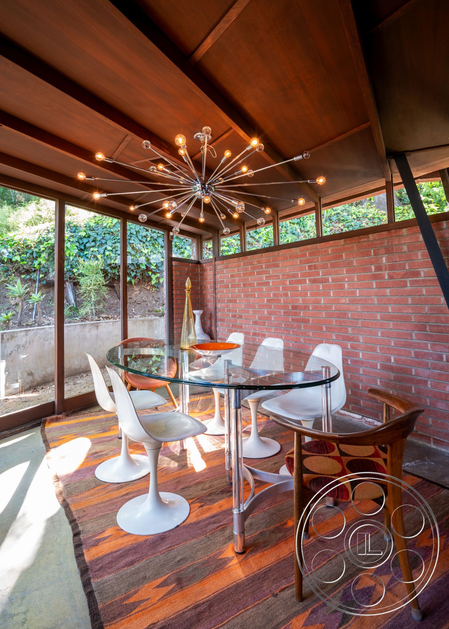 Retro 31 - floor-to-ceiling windows interior, exposed brick accent wall interior, open-concept space interior, textured area rug interior, modern lighting fixture interior, hardwood ceiling beams interior, round glass dining table interior, mid-century modern dining chairs interior, vintage patterned seating upholstery interior, bright and airy dining area interior, contemporary glass vase interior, natural light-filled dining space interior, surrounding greenery view interior