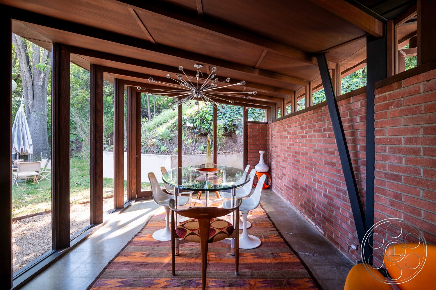 Retro 31 - natural light interior, wood beam ceiling interior, floor-to-ceiling windows interior, open floor plan interior, exposed brick accent wall interior, concrete floor interior, greenery view exterior, textured area rug interior, mid-century modern design style interior, round glass dining table interior, modern dining chairs interior, landscaped garden setting exterior, city retreat setting exterior, contemporary lighting fixture interior, smooth concrete wall interior, rustic wooden accents interior