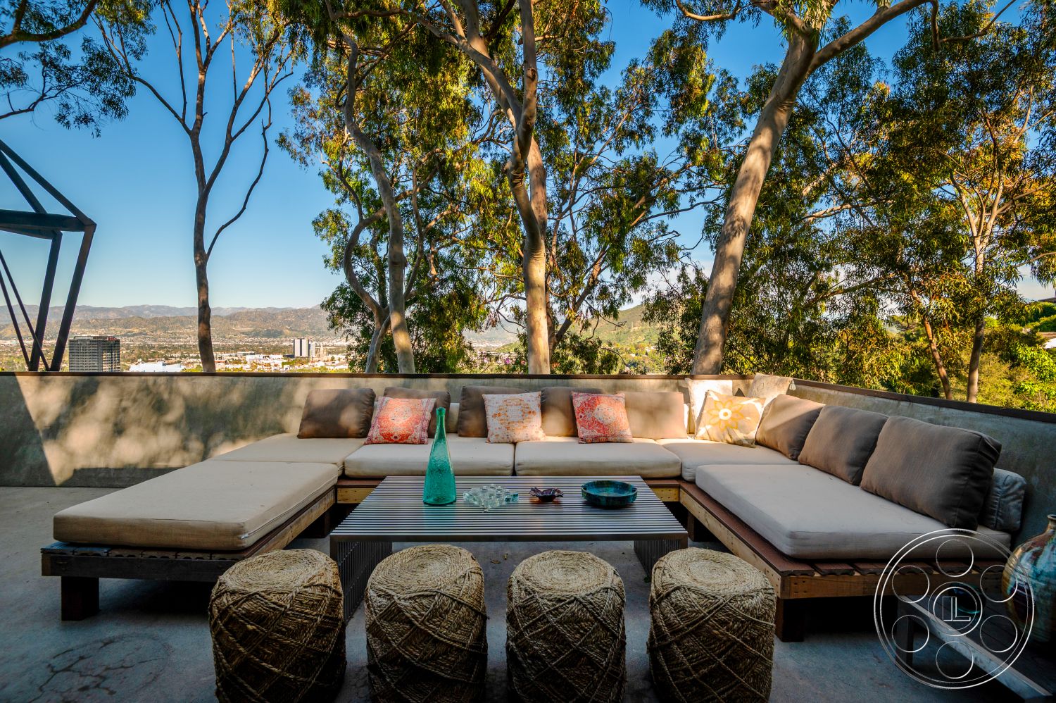 Retro 31 - minimalist decor, wooden coffee table, modern design elements, city skyline view, open-air terrace, exterior seating area, concrete patio floor, natural light ambiance, urban outdoor living space, wooden sectional sofa, upholstered outdoor cushions, straw poufs, glass water bottle, decorative serving tray, outdoor container plants, surrounding eucalyptus trees, landscape orientation