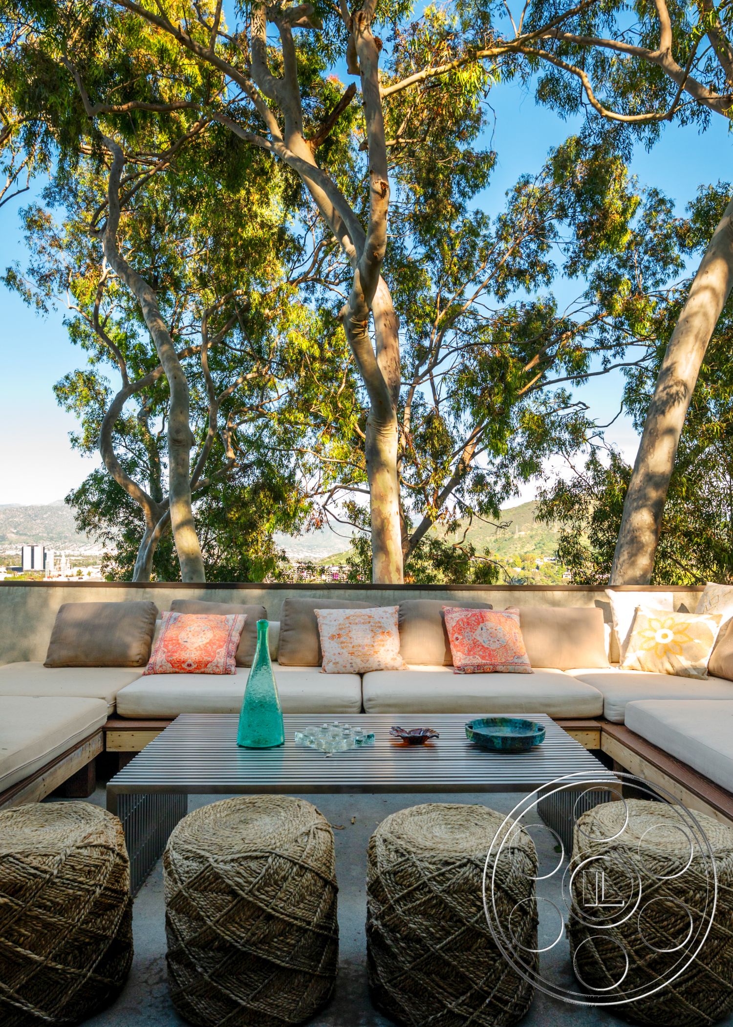 Retro 31 - urban skyline view, decorative throw pillows, open-air setting, modern outdoor design, relaxed outdoor living space, soft fabric outdoor cushions, exterior lounge area, natural landscape integration, wooden seating platform, concrete table surface, woven straw stools, sustainable bamboo coffee table, glass vase centerpiece, geometric glass tableware, lush eucalyptus trees surrounding