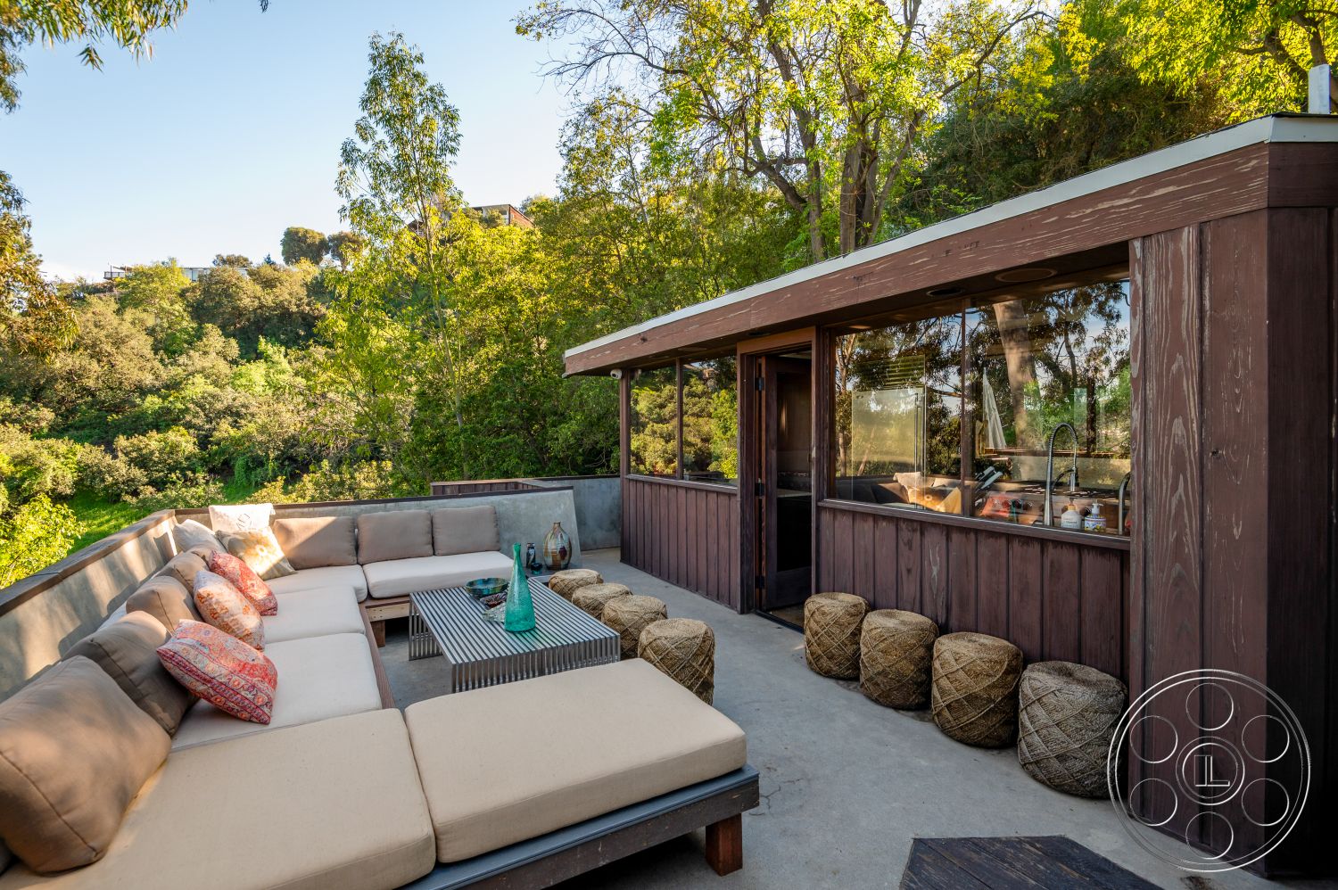 Retro 31 - exterior, glass sliding doors, open space design, wooden deck, natural light from large windows, dark wood paneling, woven poufs, urban hillside location, landscaped garden setting, concrete patio floor, contemporary outdoor living area, modern sectional outdoor seating, organic material finishes, textured rug on patio, wooden ceiling with exposed beams, rustic outdoor kitchen area, integrated seating and table structure, comfortable outdoor entertaining area