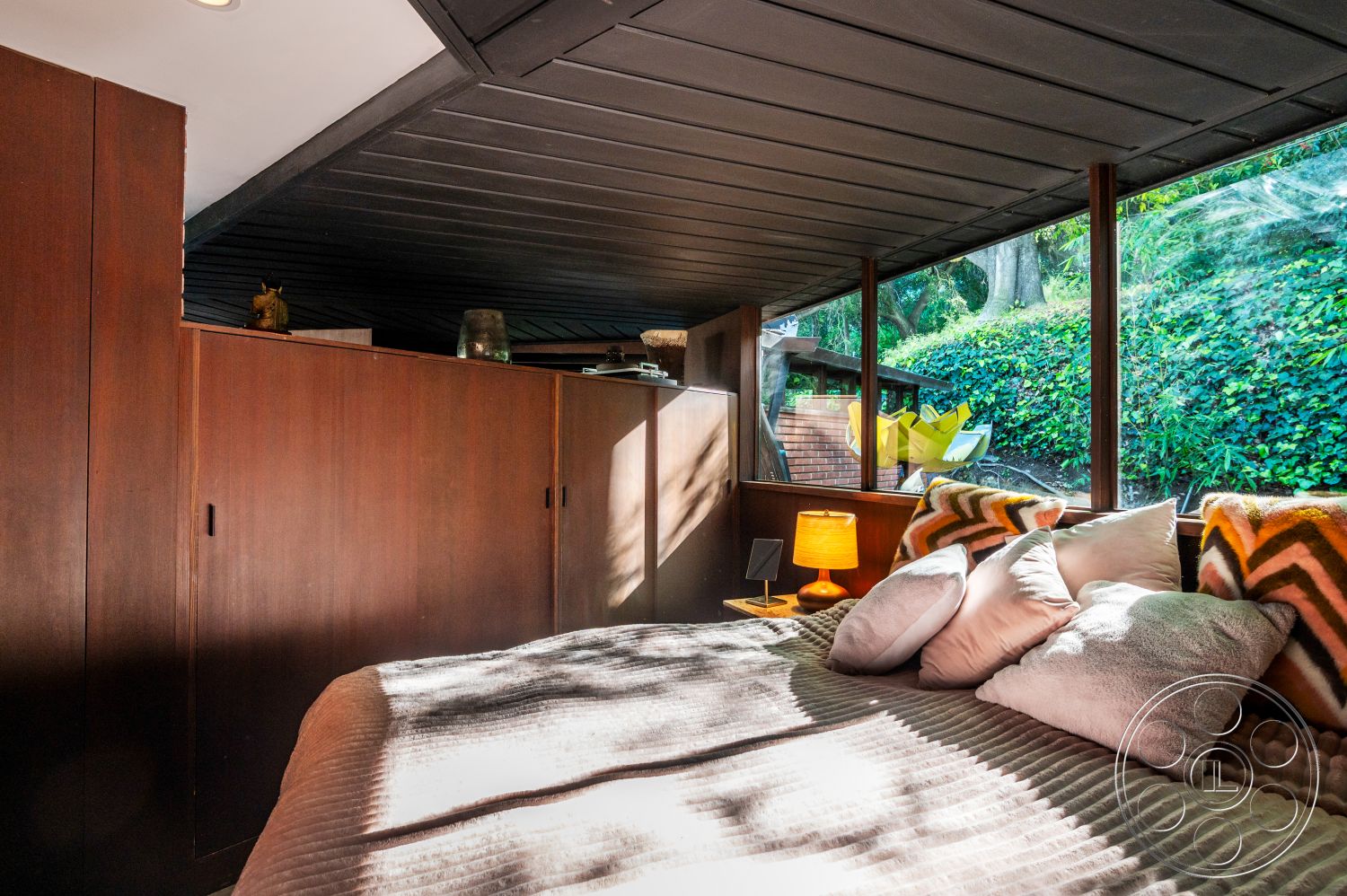 Retro 31 - minimalist decor, indoor-outdoor flow, textured throw pillows, cozy reading nook, earth-toned color palette, contemporary interior design, decorative accent pieces, clutter-free space, natural light interior, serene forest setting, vaulted ceiling with wood paneling, hardwood bedroom walls, large glass windows overlooking greenery, modern bedroom lighting, layered bedding textures, exposed wood storage cabinetry, patterned throw blankets, L-shaped bedroom layout, wood furniture finishes