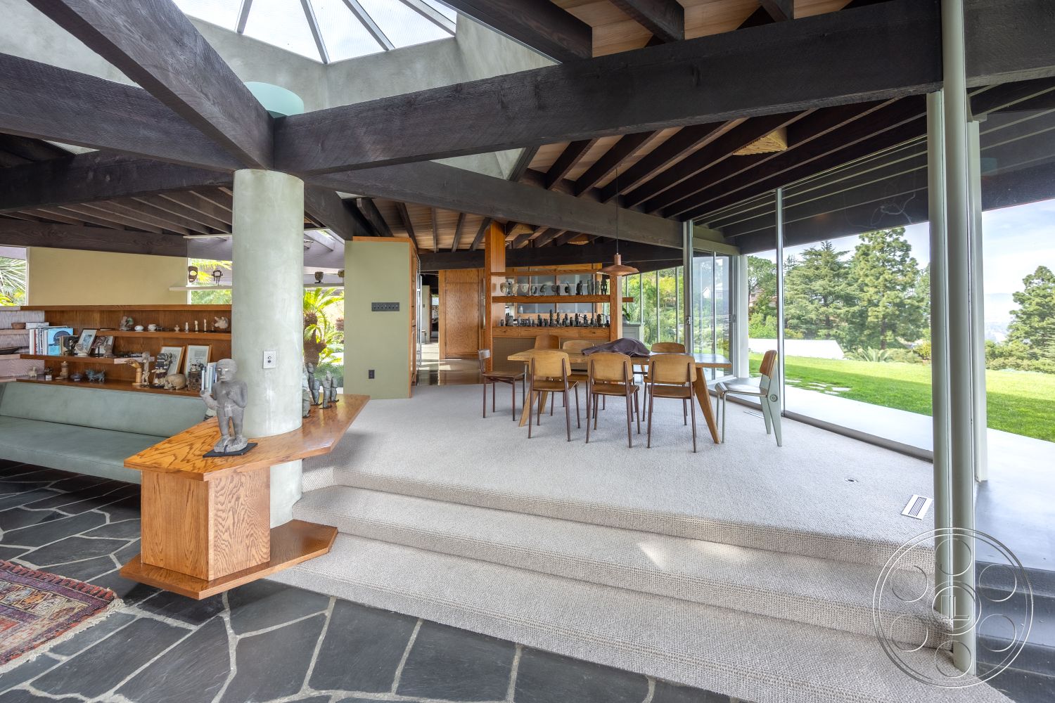 Retro 19 - open floor plan, slate stone flooring, interior space, decorative art pieces, natural wood dining table, textured wall finishes, seamless indoor-outdoor flow, contemporary lighting fixtures, mid-century modern design, exposed wooden ceiling beams, glass wall with garden view, built-in wooden shelving unit, carpeted living room steps, soft neutral carpet, outdoor landscaping visible, polished concrete columns