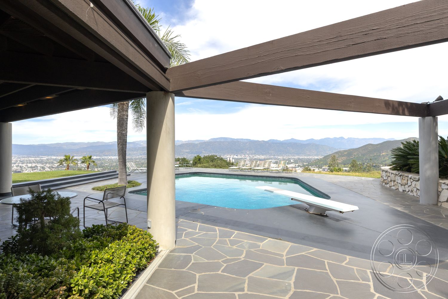 Retro 19 - recessed lighting, exterior, modern design elements, glass railing, city skyline view, mountain backdrop, open-air patio, natural stone wall, rectangular pool, exposed wood ceiling beams, landscaped garden area, stone patio flooring, concrete pool border, integrated lighting features, stainless steel pool ladder