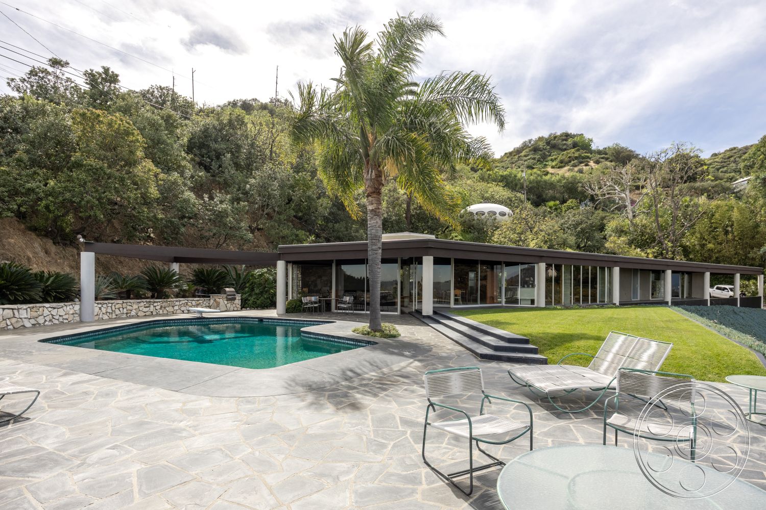 Retro 19 - exterior, palm trees, minimalist design elements, earthy color palette, glass sliding doors, low-maintenance garden, rectangular swimming pool, tropical landscaping, poolside lounge chairs, hillside location, exposed stone wall, contemporary home design, concrete pool border, natural stone patio flooring, framed glass windows, flat roof architecture, modern outdoor living space, landscaped green lawn