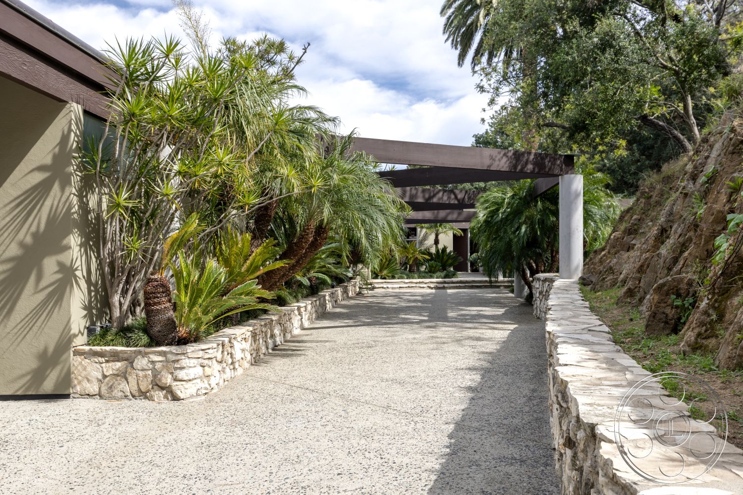 Retro 19 - exterior concrete driveway, exterior hillside location, exterior muted color palette, exterior tropical landscaping, exterior stone retaining wall, exterior natural light, exterior aggregate concrete surface, exterior stone border wall, exterior wood pergola structure, exterior palm tree accents, exterior modern architecture details, exterior organic forms