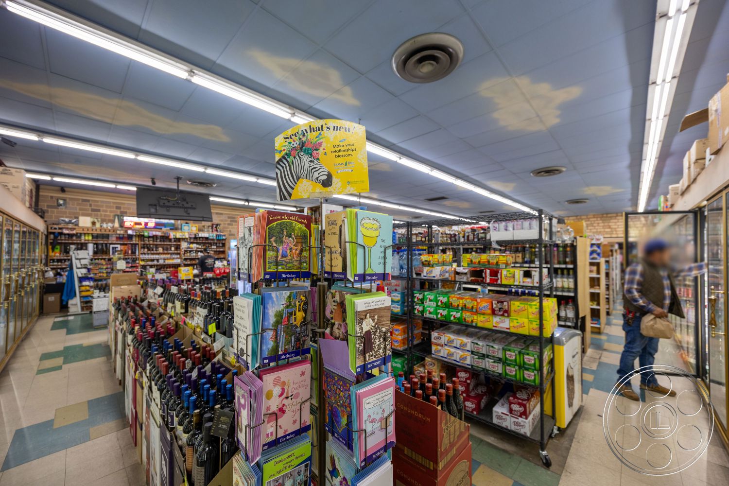 Shop 128 - fluorescent ceiling lights, colorful product packaging, cardboard product displays, organized shelving units, glass refrigerator doors, grocery store aisles, blue tiled ceiling, wooden display shelves, linoleum floor tiles, green and beige checkered tile flooring, variety of wine bottles, snack food displays, beverage coolers, merchandise signage