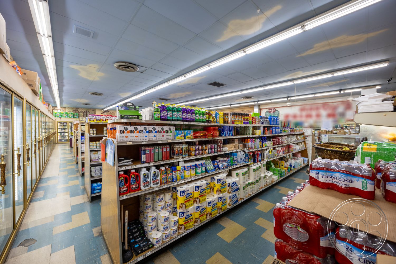 Shop 128 - wooden shelves, fluorescent ceiling lights, vinyl tile flooring, blue drop ceiling, checkered blue and beige floor tiles, refrigerated glass display cases, variety of product packaging, shelves stocked with household goods, clean and organized aisles, well-lit shopping environment, grocery store layout
