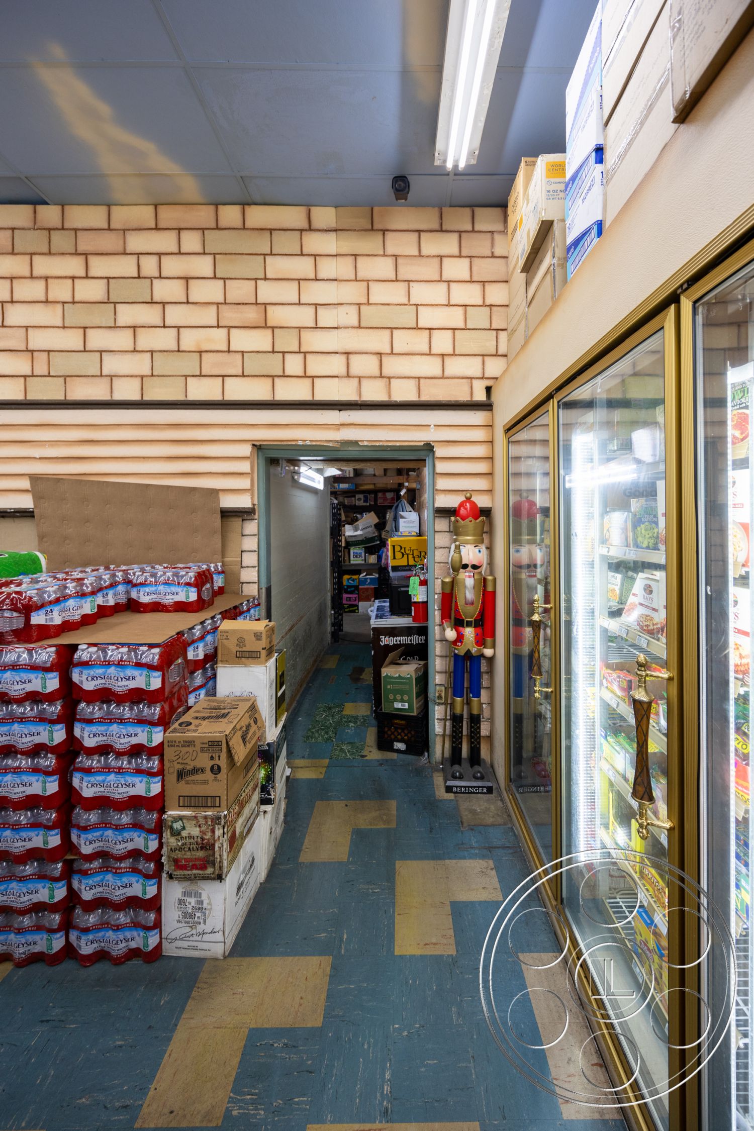 Shop 128 - vinyl flooring with blue and yellow patterns, exposed ceiling with fluorescent lighting, textured wall with beige brick finish, shelving display with beverage boxes, glass refrigerator display with metal frame, cardboard packaging for products, nutcracker decoration, narrow store aisle layout