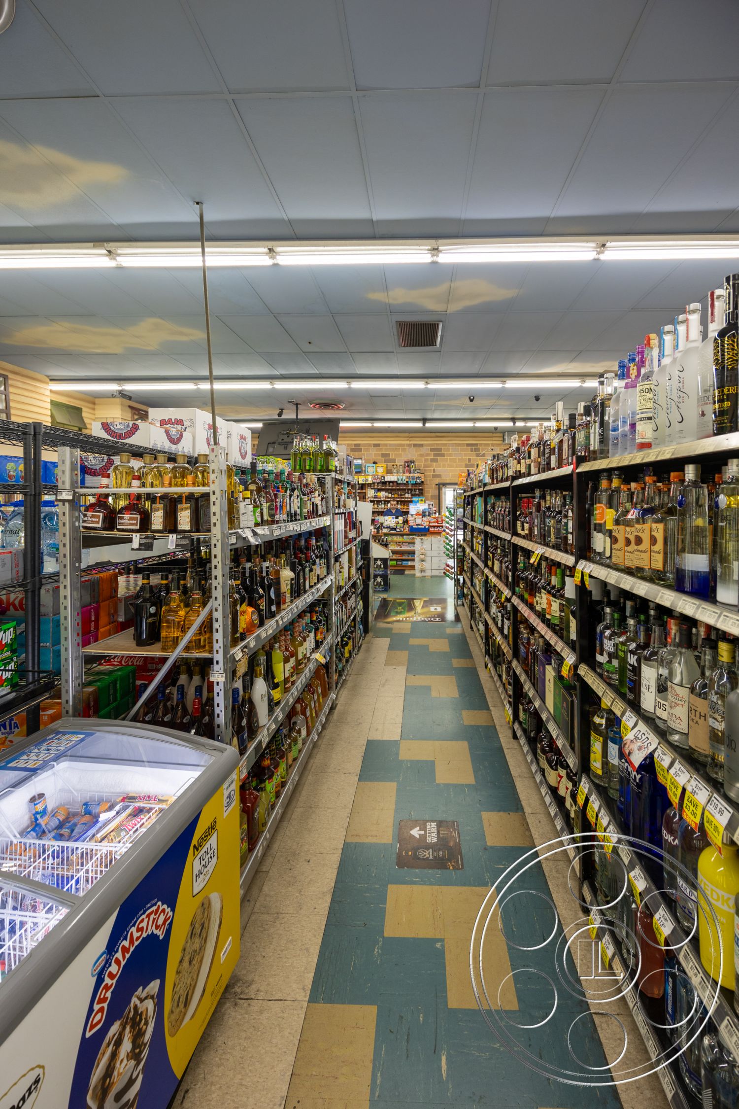Shop 128 - drop ceiling tiles, wide aisle layout, linoleum flooring, blue and yellow tile floor, commercial shelving units, beverage display shelves, refrigerated ice cream freezer, ambient ceiling lights, brick wall construction