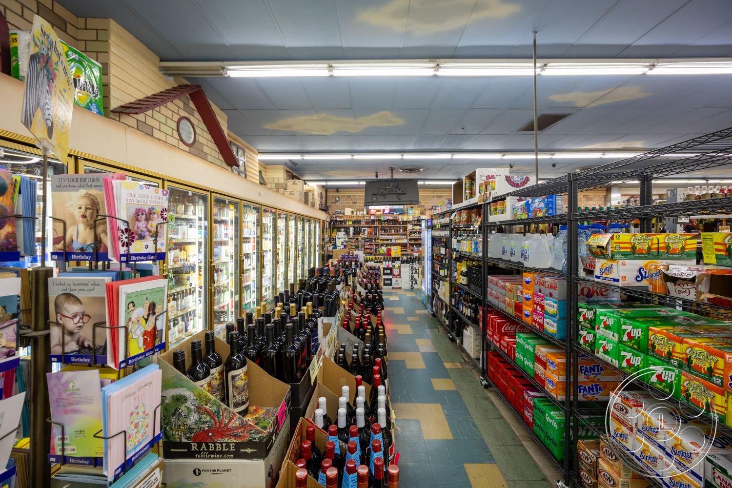 Shop 128 - strip lighting, checkerboard tile flooring, interior convenience store, glass refrigerator doors, brick wall accent, shelf display for beverages, bottled wine display, colorful snack packaging, checkout cards stand, light blue ceiling tiles, variety soft drink boxes, card display rack, beverage shelving units, snack aisle