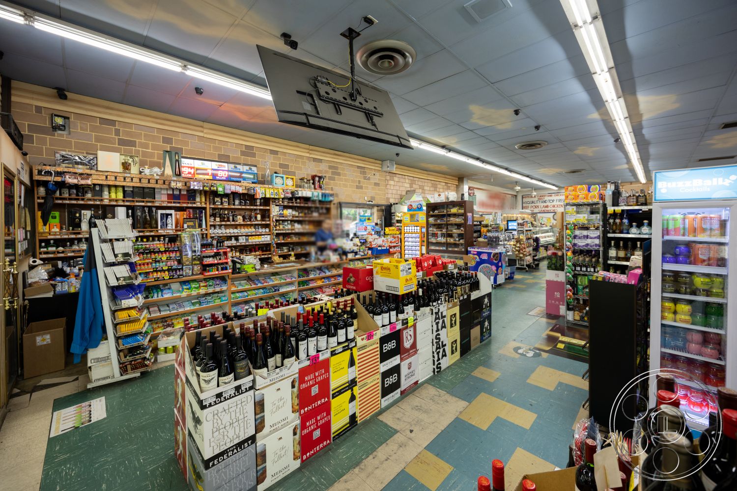 Shop 128 - brick accent wall, wooden shelving units, fluorescent ceiling lights, vinyl tile flooring, refrigerated display cases, colorful product packaging, checkout counter area, shelves filled with products, aisles with wine displays, variety of beverage options, general store layout