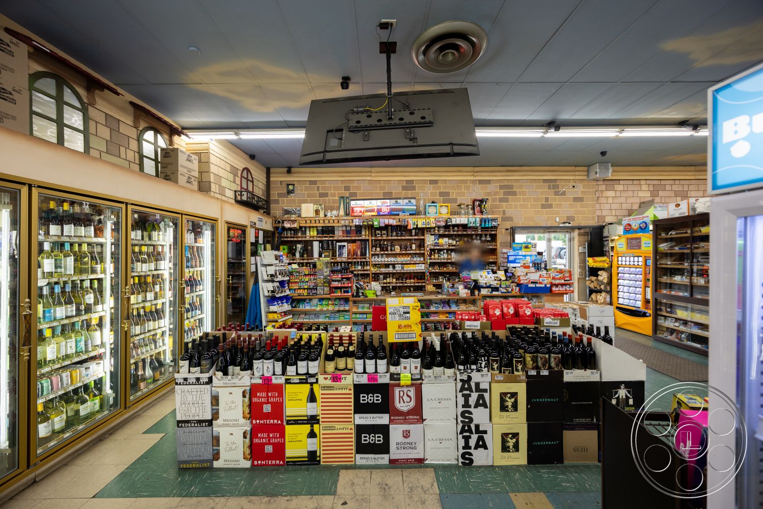 Shop 128 - exposed brick wall, suspended ceiling, glossy tile flooring, LED lighting fixtures, open shelving units, colorful packaging, case display refrigerators, rows of wine bottles, wooden product displays, green tile flooring, retail checkout area