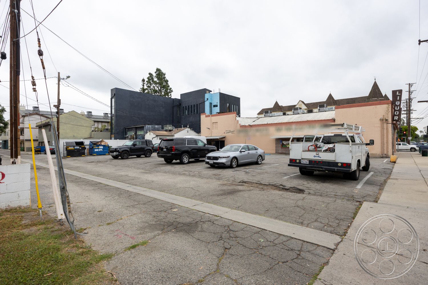 Shop 128 - exterior, asphalt driveway, wooden fence, red brick walls, concrete parking lot, blue dumpsters, adjacent residential buildings, gray asphalt surface, modern flat-roof building, white painted signage, metal pole supporting power lines, commercial building with large windows, angled rooftop, vintage liquor sign, surrounding urban setting