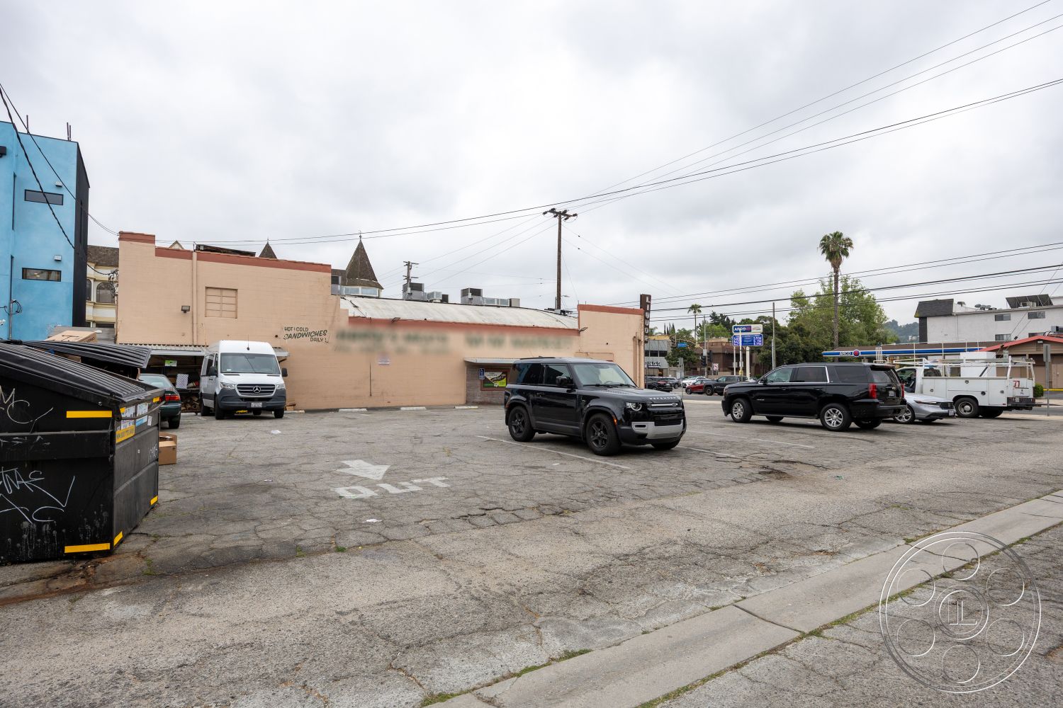 Shop 128 - exterior, paved driveway, asphalt parking lot, surrounding urban environment, commercial building facade, painted concrete walls, overcast sky, palm trees in urban setting, flat roof building, telephone utility poles, mixed-use commercial area, dumpster with graffiti
