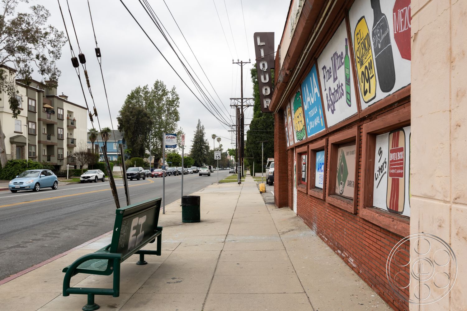 Shop 128 - green landscaping, concrete sidewalk, urban commercial setting, painted signage, overcast sky, brick storefront facade, utility pole, paved roadway, pedestrian bench, multifamily residential buildings, public transportation signage