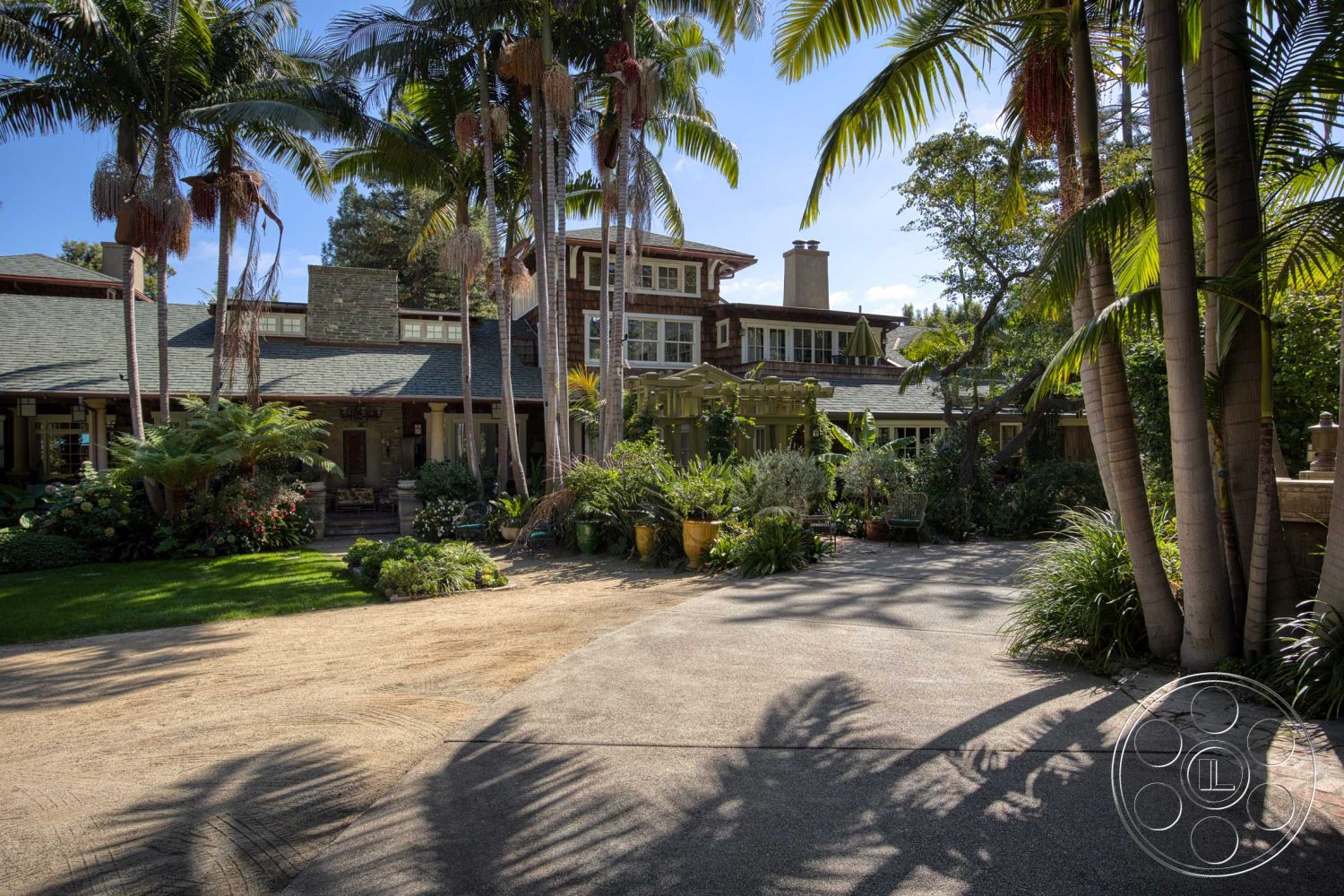 Americana 48 - exterior, lush greenery, palm trees, tropical landscaping, stucco walls, garden patio, modern farmhouse style, cobblestone driveway, shingle roof, stone chimney, two-story home, sloped roof design, covered entryway, decorative flower pots, wooden porch railing, multisided windows