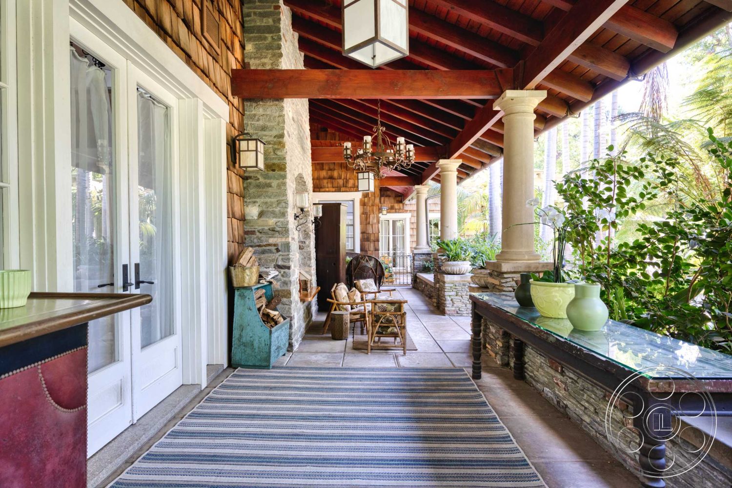 Americana 48 - exterior, stone accent wall, transom windows, double French doors, arched entryway, lush tropical garden, wood beam ceiling, rustic wood furniture, natural light-filled area, built-in stone seating, shingle wall cladding, rectangular patio flooring, tile porch flooring, decorative hanging lantern, outdoor chandelier