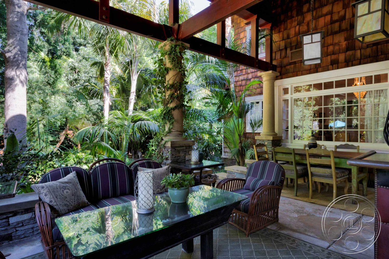 Americana 48 - exterior, glass table top, exposed wood beam ceiling, rustic wooden dining table, natural stone patio flooring, tropical garden setting, stone column supports, wood shingle siding, wicker seating with striped cushions, large windows with grid pattern, lush greenery and palm trees