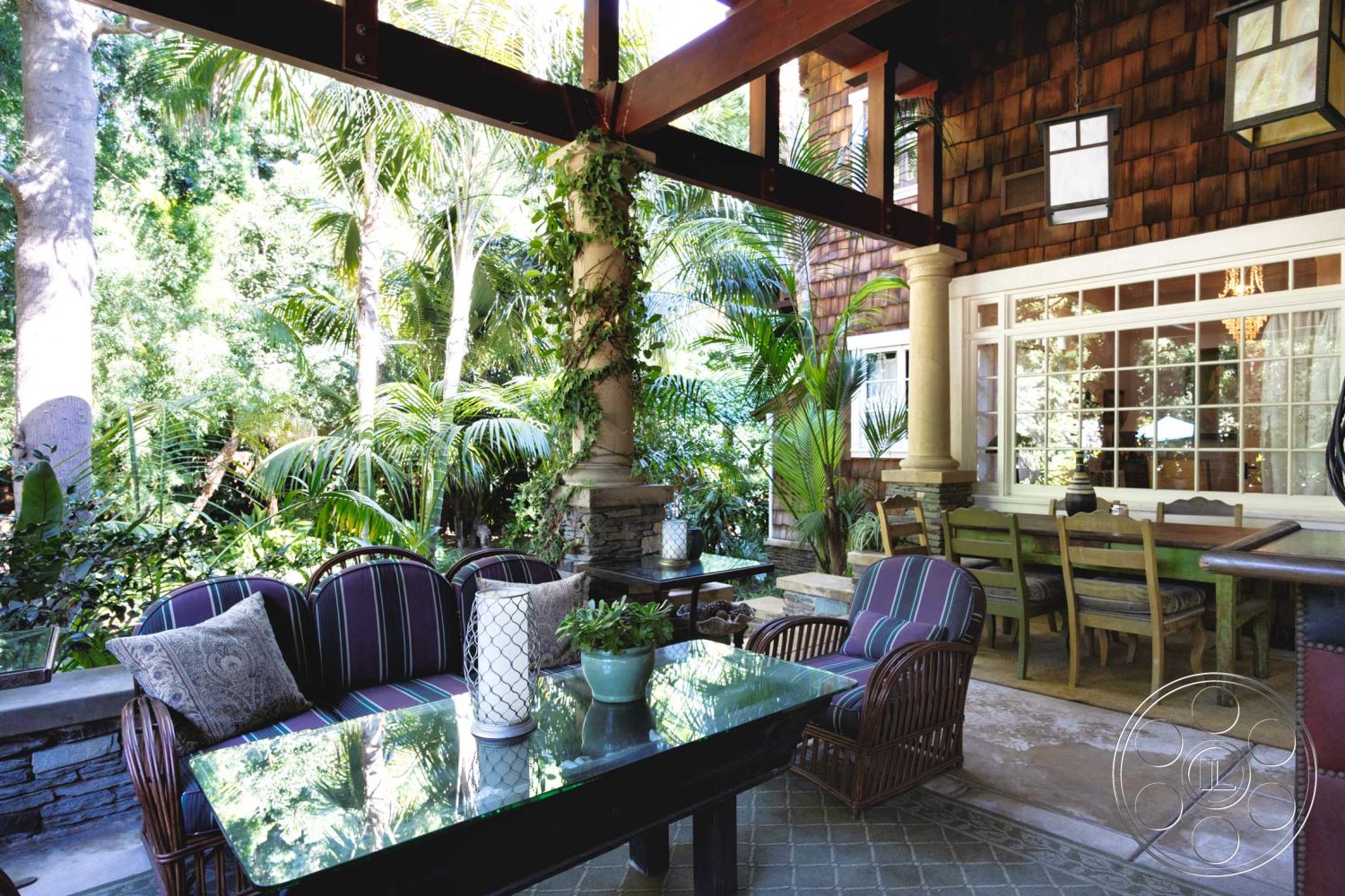 Americana 48 - exterior, wooden beam ceiling, decorative potted plants, natural stone columns, slate patio flooring, woven rattan seating, dining area with wooden table, tropical garden setting, large windows with grid pattern, shingled accent wall, glass top patio table, courtyard with lush foliage