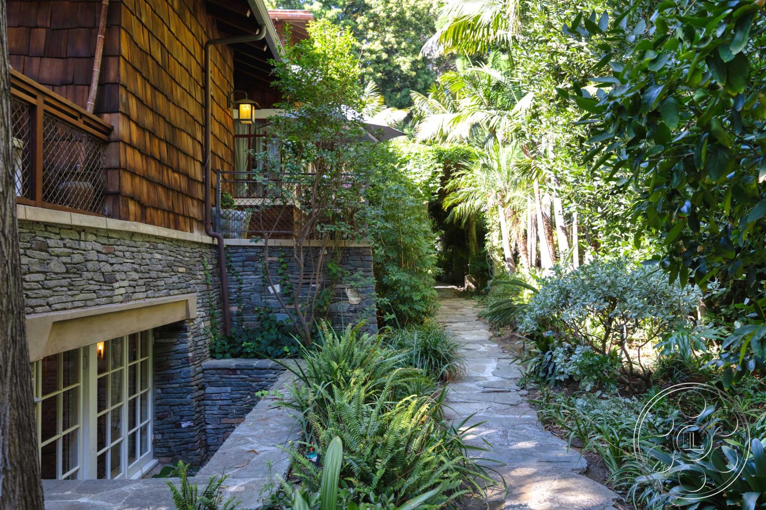 Americana 48 - exterior, large glass windows, tropical foliage, natural stone texture, tall palm trees, flagstone pathway, lush garden landscaping, metal balcony railing, wood beam details, varied shrubbery, wood shingle siding, stacked stone wall, exposed stone foundation