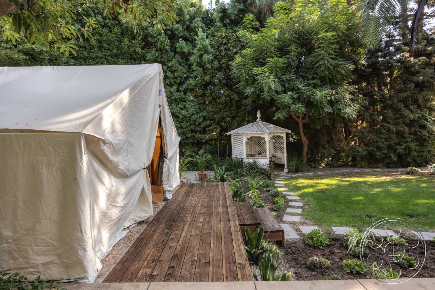 Americana 48 - lush greenery, stone stepping stones, patio seating area, landscaped garden area, natural stone border, exterior garden setting, wooden deck pathway, flat lawn area, canvas tent exterior, white gazebo structure